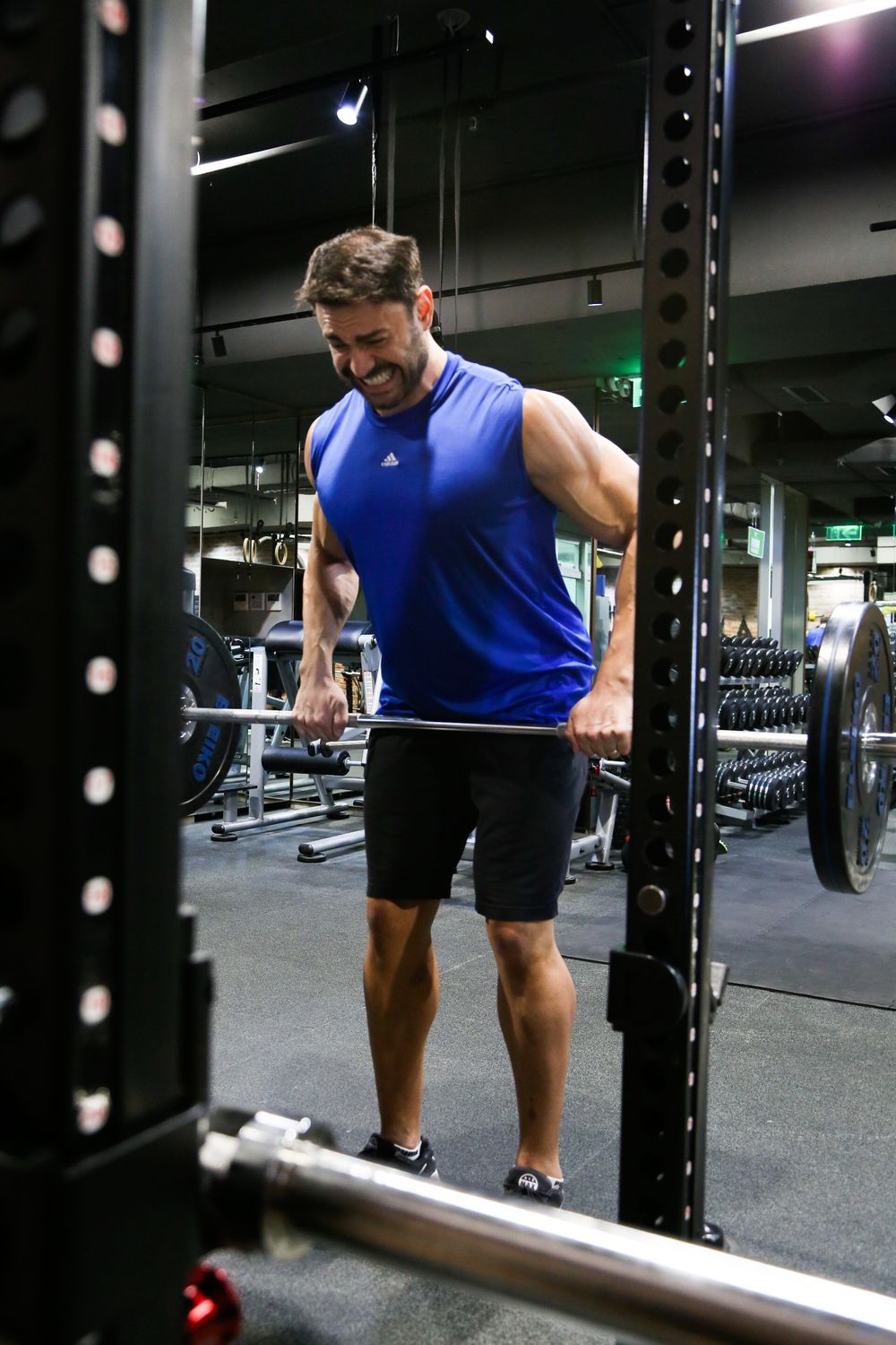 Muscular man in blue Adidas sleeveless shirt doing barbell exercise at modern gym.