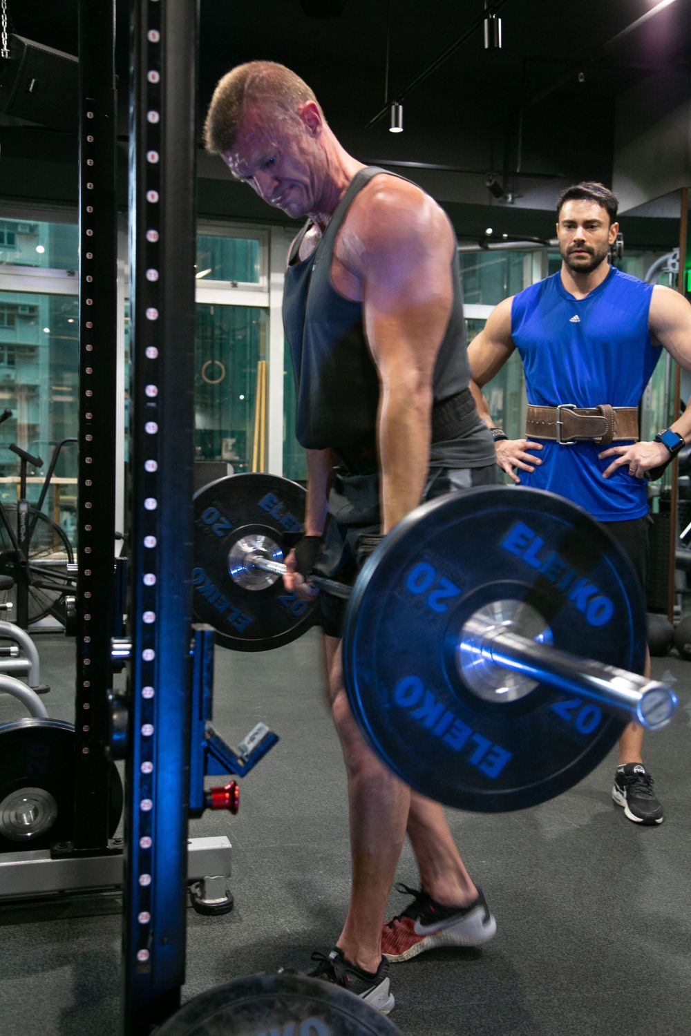 Two muscular men training with heavy barbell weights at a modern gym power rack.