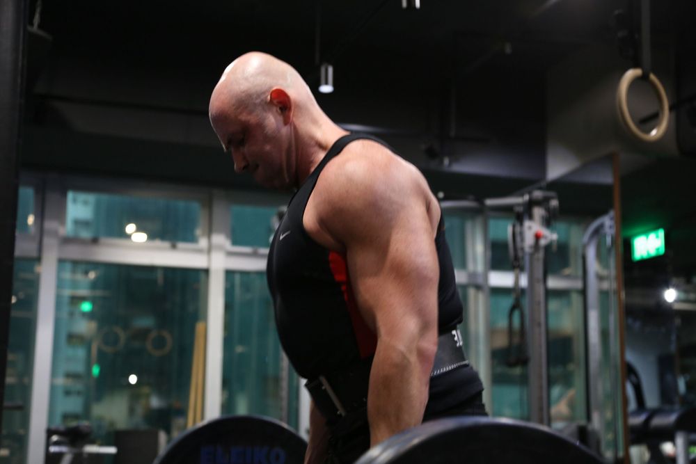 Bald bodybuilder in black tank top bent over barbell rows showing massive back muscles in gym.