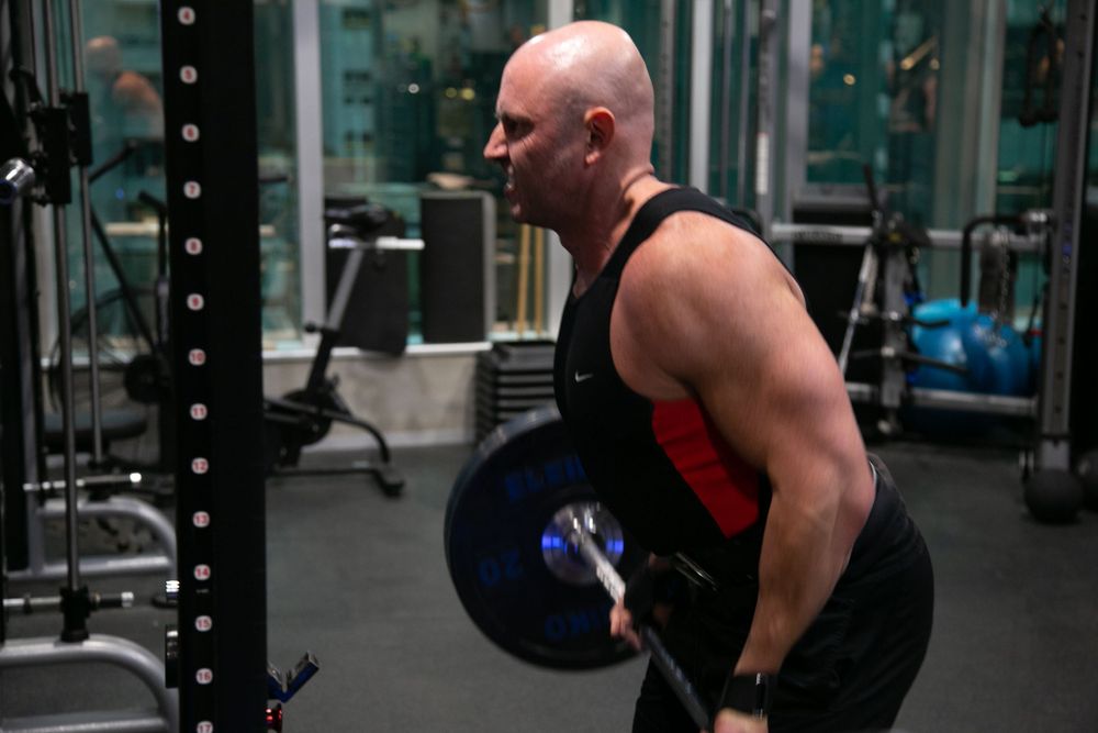 Bald muscular man in black and red tank top performing bent-over barbell rows in dark gym.