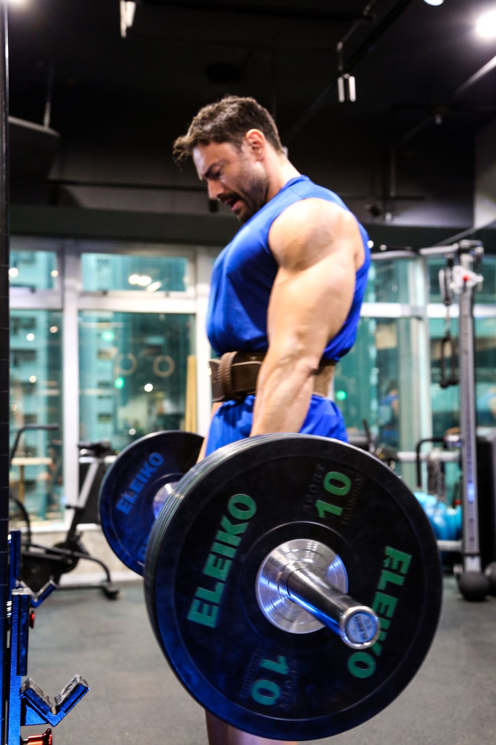 Muscular man in blue sleeveless shirt lifting heavy Eleiko barbell in bent-over row position.