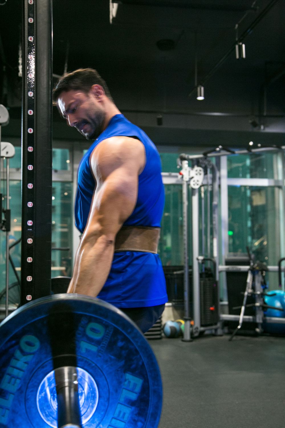 Athletic man in blue tank top performing bent-over barbell row with Eleiko weights in gym.