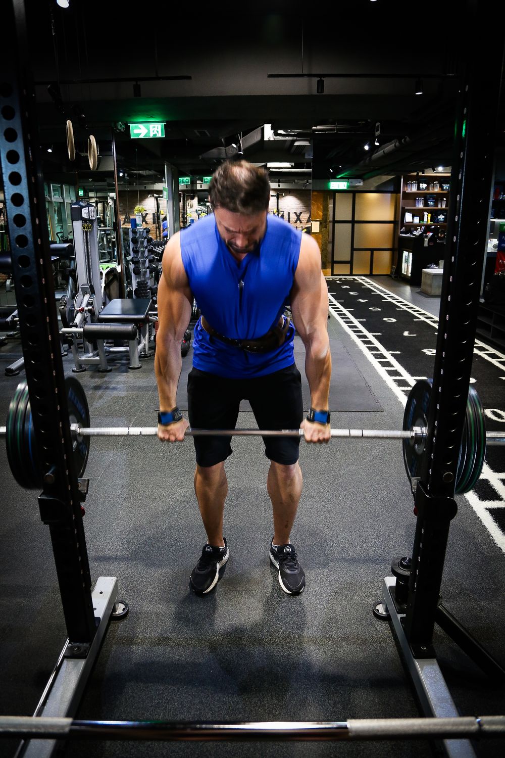 Muscular man in blue sleeveless shirt performing bent-over barbell rows in well-equipped gym.