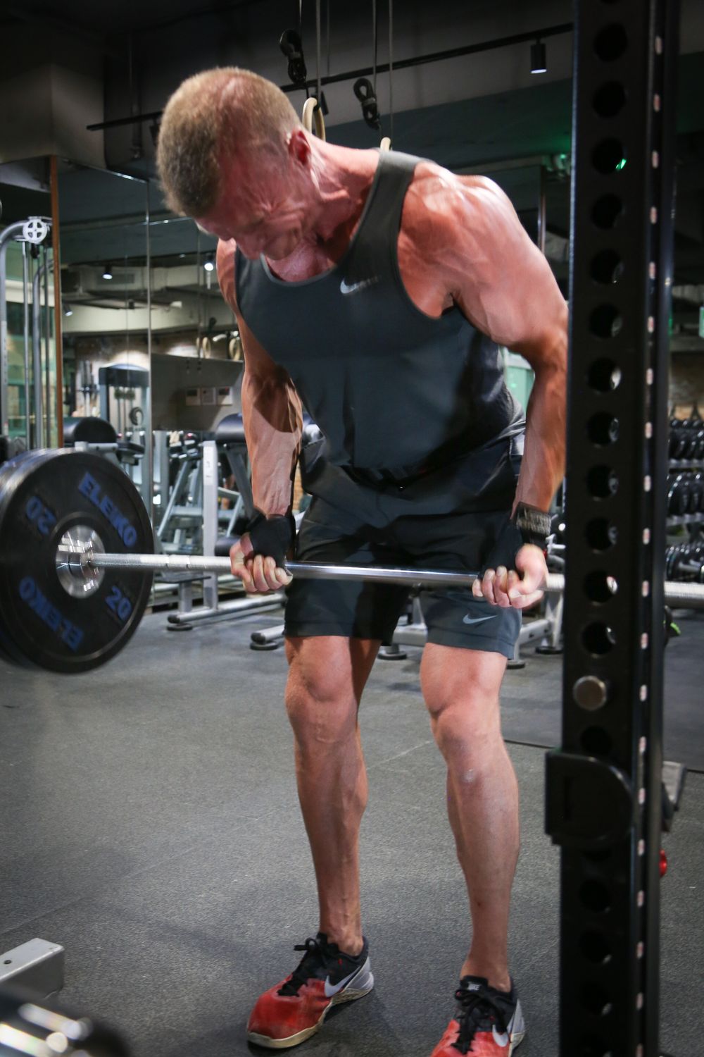 Lean older athlete in Nike gear performing bent-over barbell rows with heavy weights in gym.