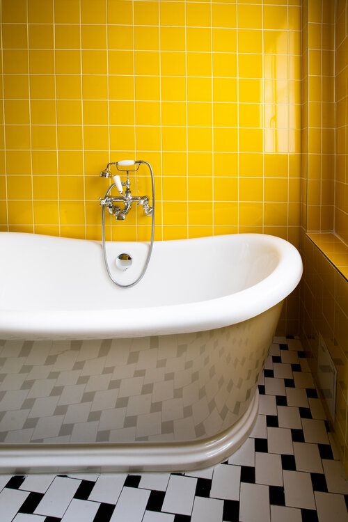 Vintage clawfoot bathtub against bright yellow tiled wall with black and white floor.
