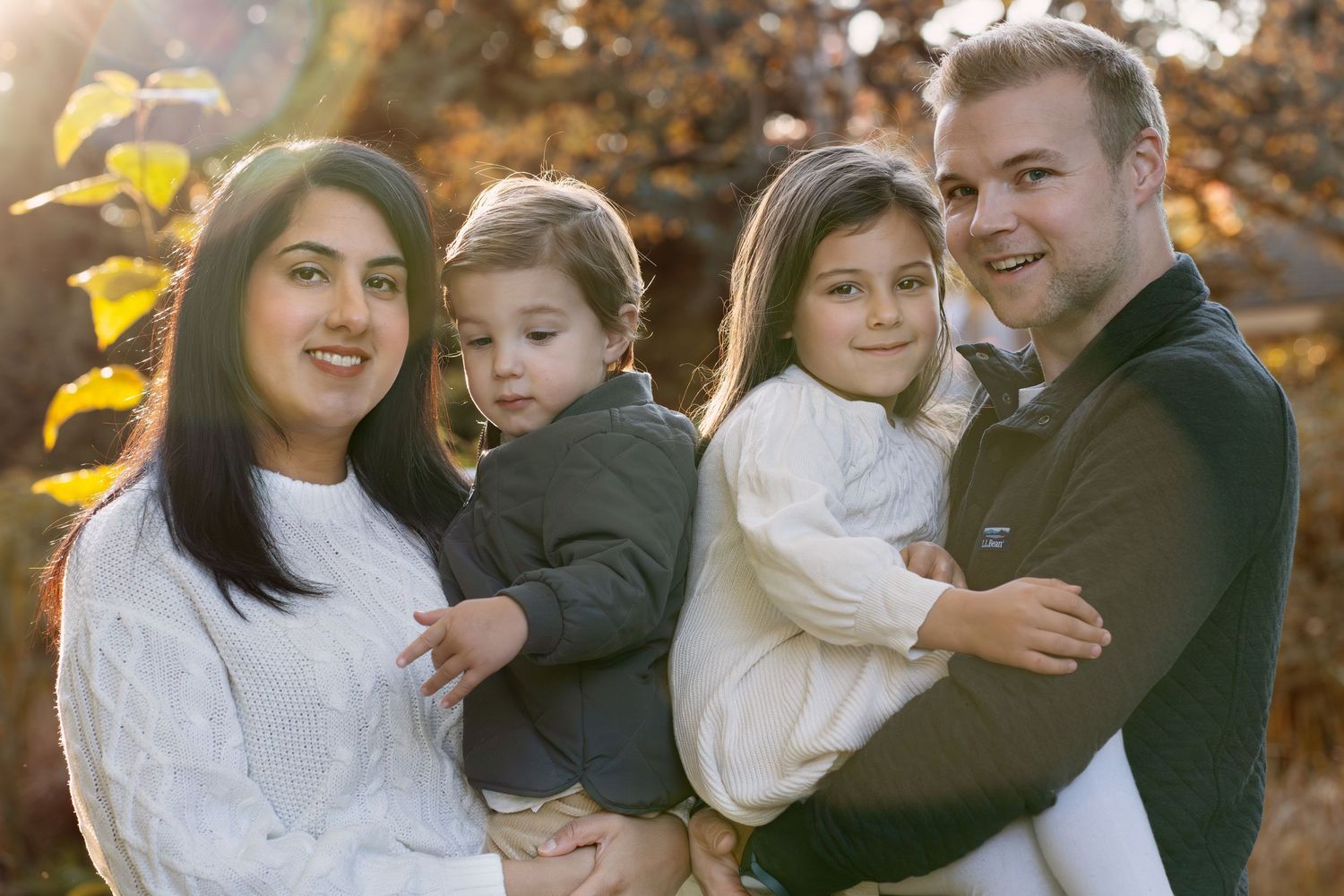 A family of four poses together outdoors on an autumn day wearing coordinated white and dark colored outfits.