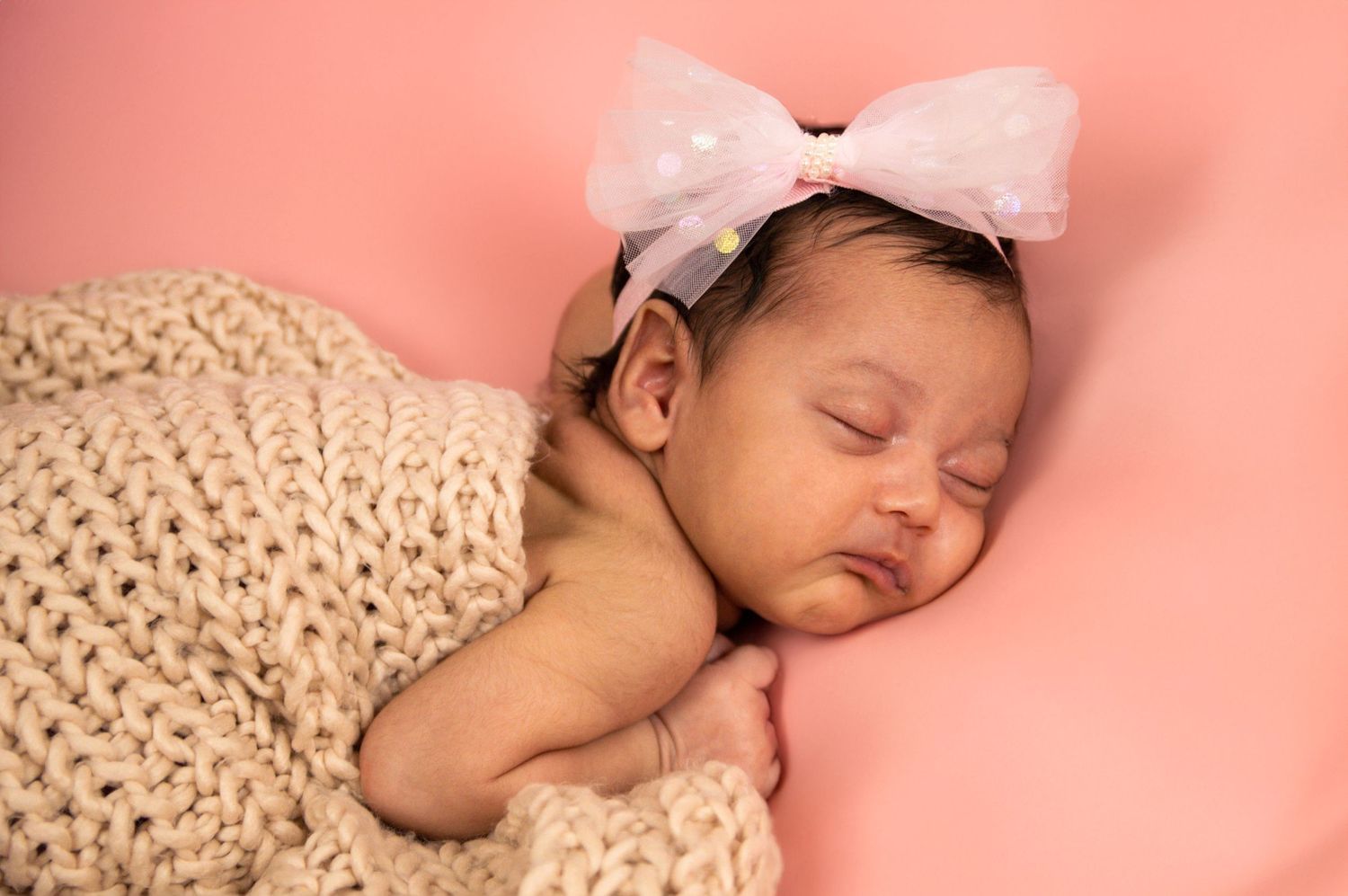 Sleeping newborn baby wrapped in knit blanket against pink background.