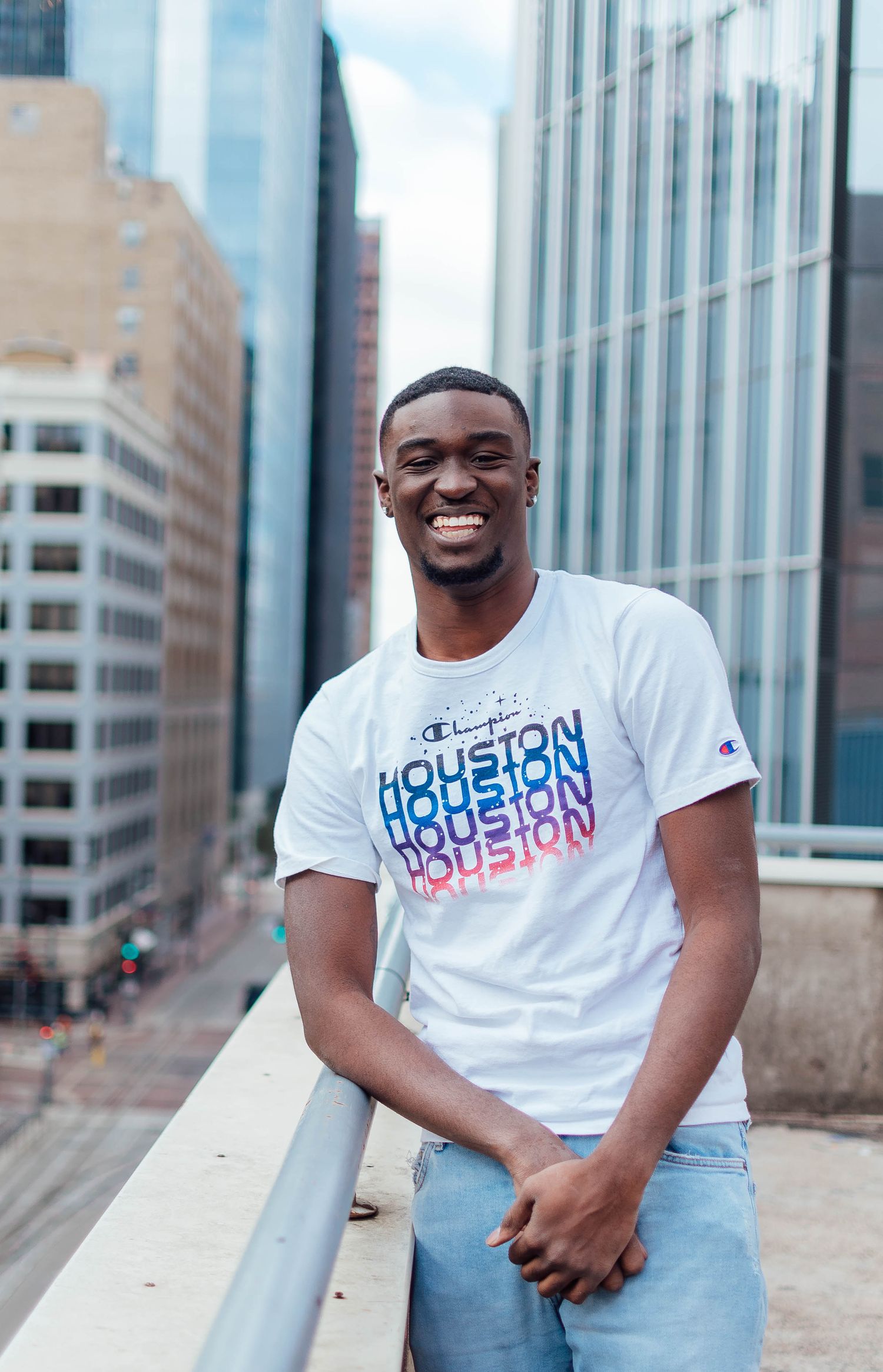 Young man in Houston t-shirt smiles against urban backdrop of downtown buildings.