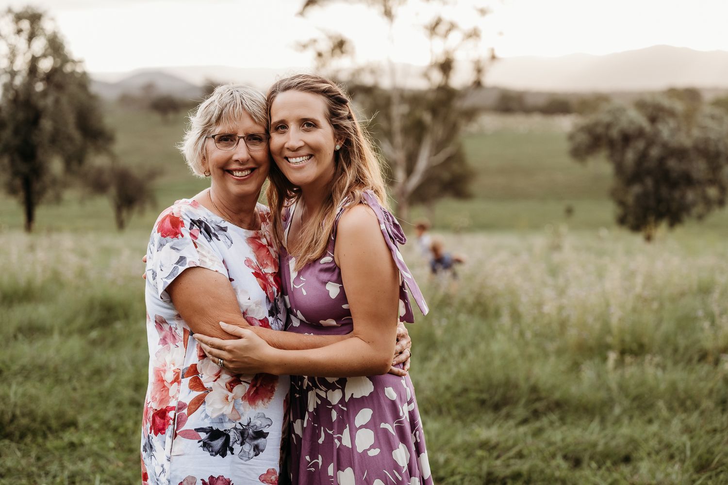 Two generations embrace in flowered dresses against a rural landscape backdrop.