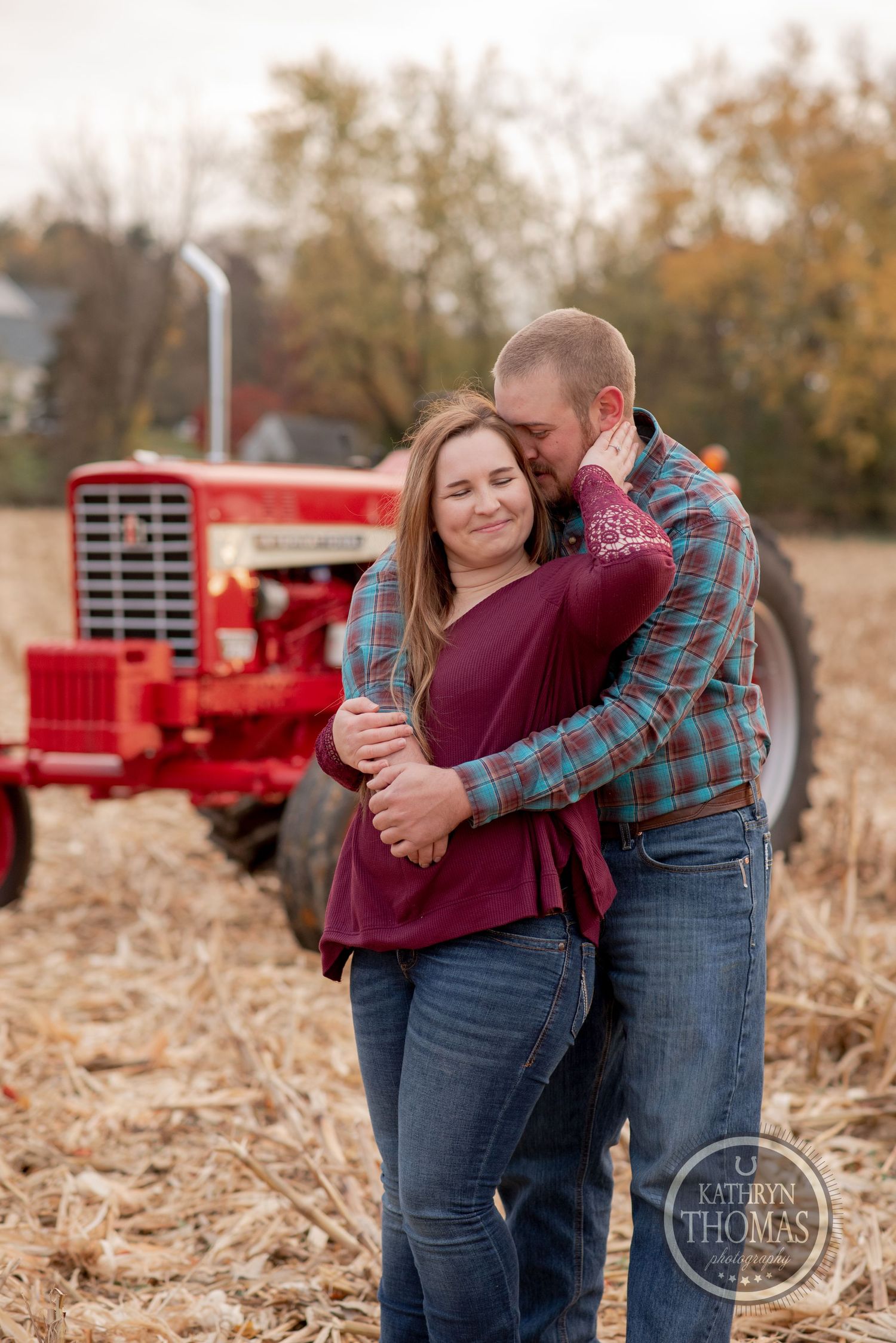 A couple embraces in front of a red tractor in an autumn cornfield.