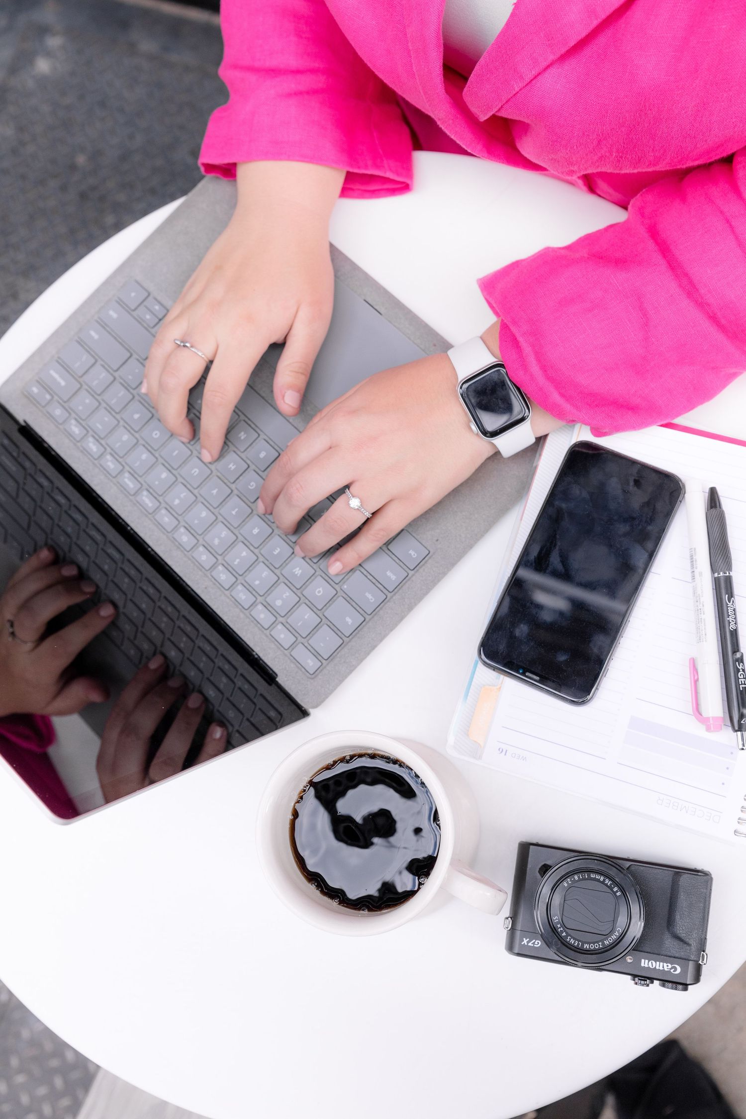 A woman in a pink blazer types on a laptop at a white table with coffee and a camera nearby.