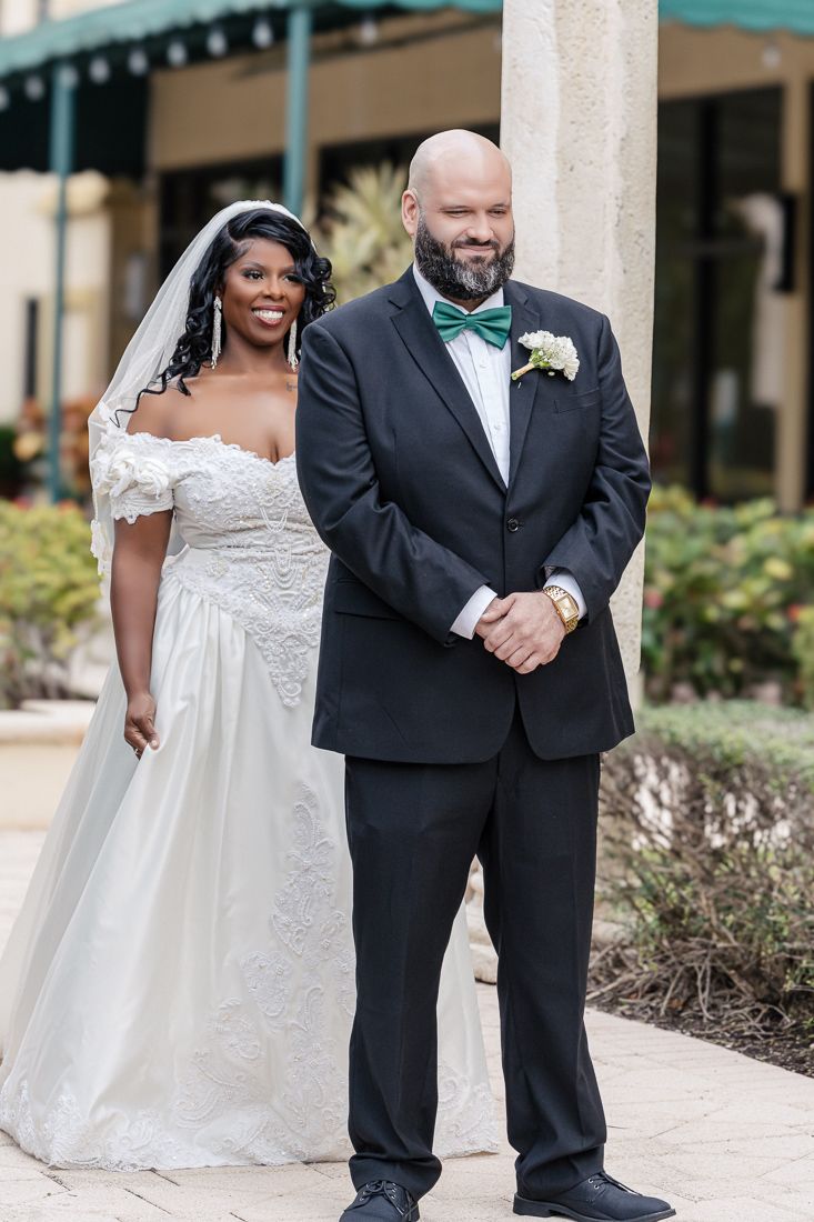 Newlywed couple poses together outdoors, bride in white lace gown and groom in tuxedo with teal bow tie.