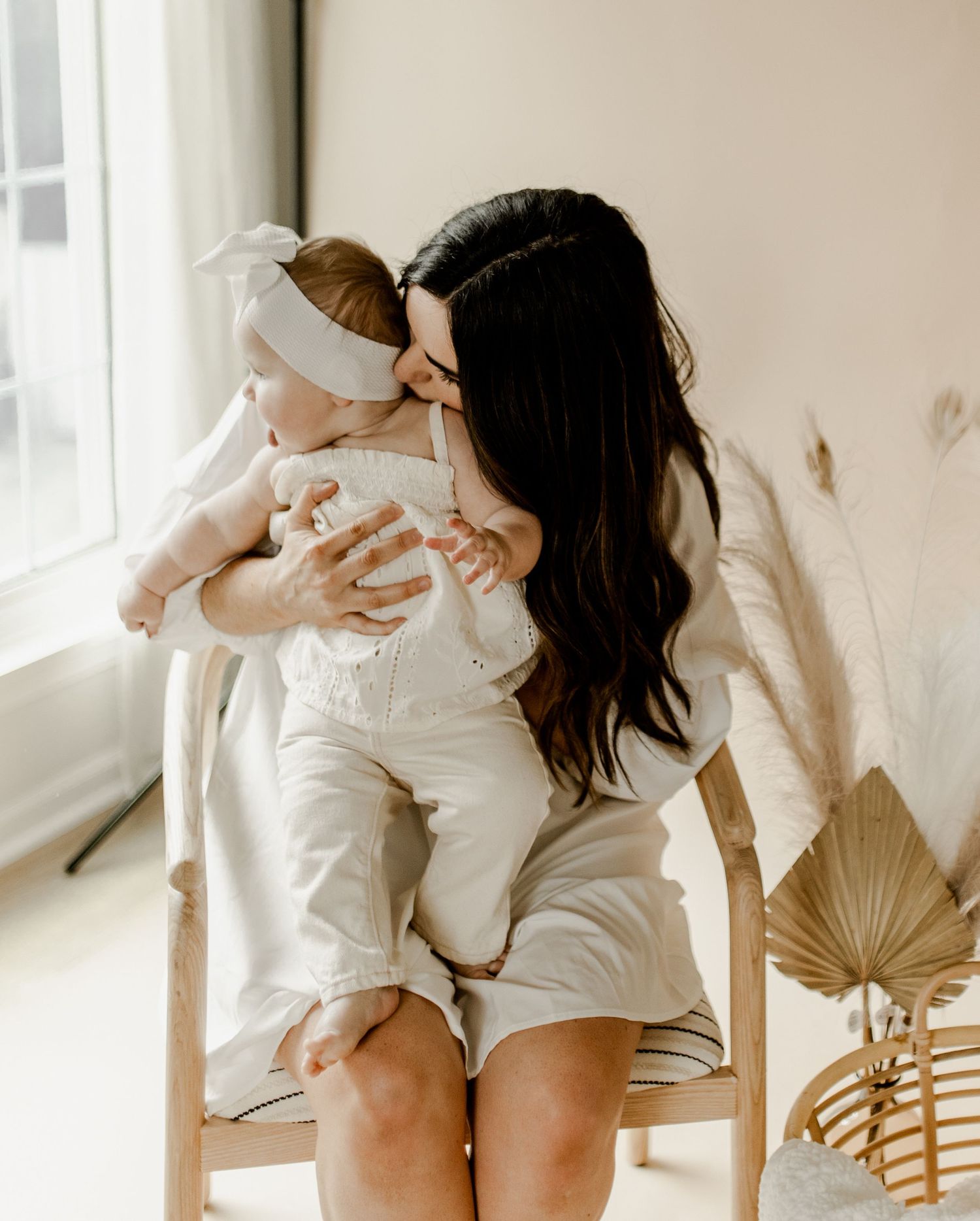 Intimate indoor portrait of mother and baby sitting by window in white clothing.