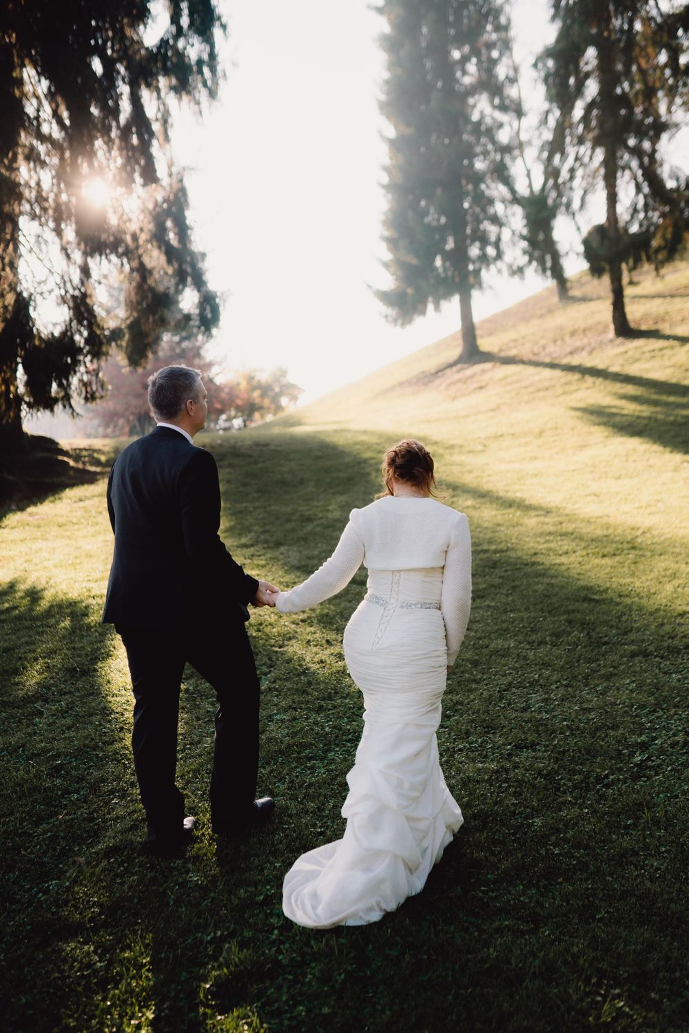 Wedding couple walking up grassy hill during sunset with evergreen trees in background.