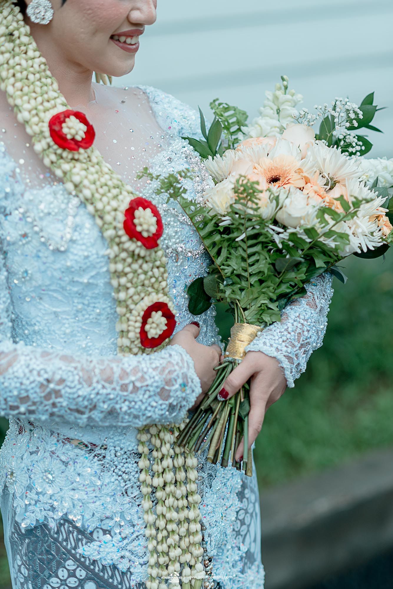 Traditional bride in lace dress holds elegant bouquet with decorative floral garland and red accents.