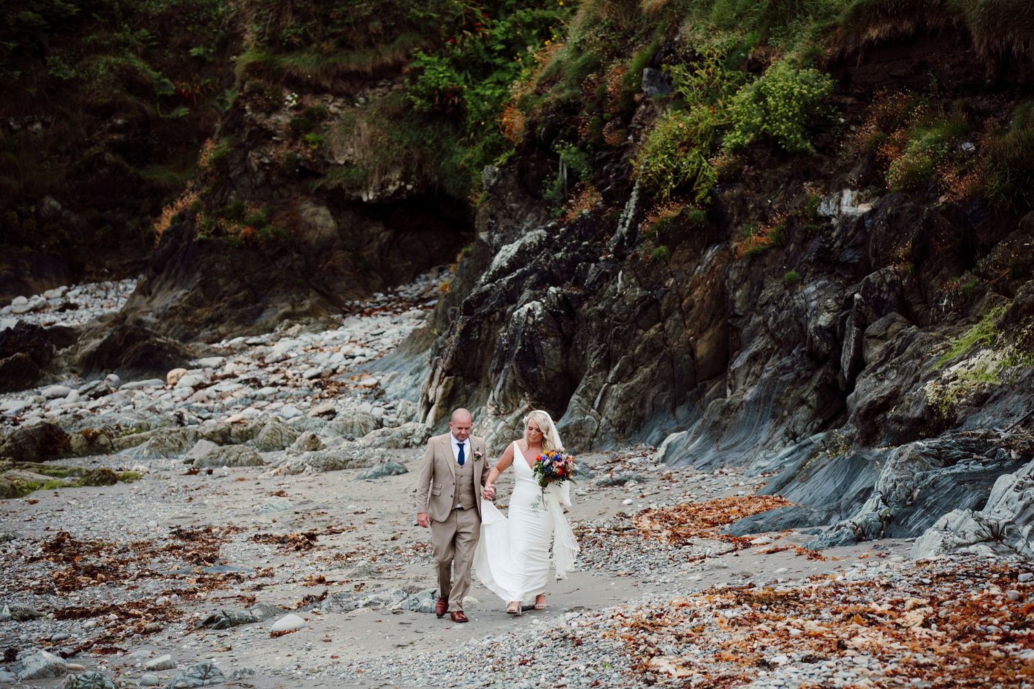 A wedding couple walks along a rocky beach shore with dramatic cliffs in the background.