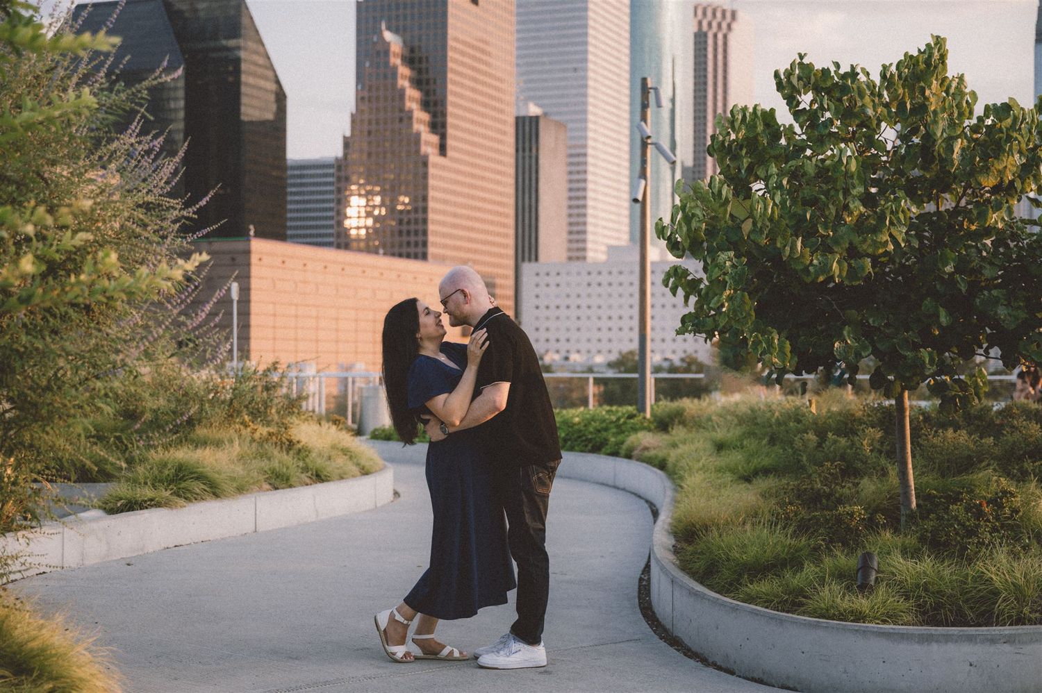 A couple embraces on a curved pathway with downtown Houston skyline in the background.