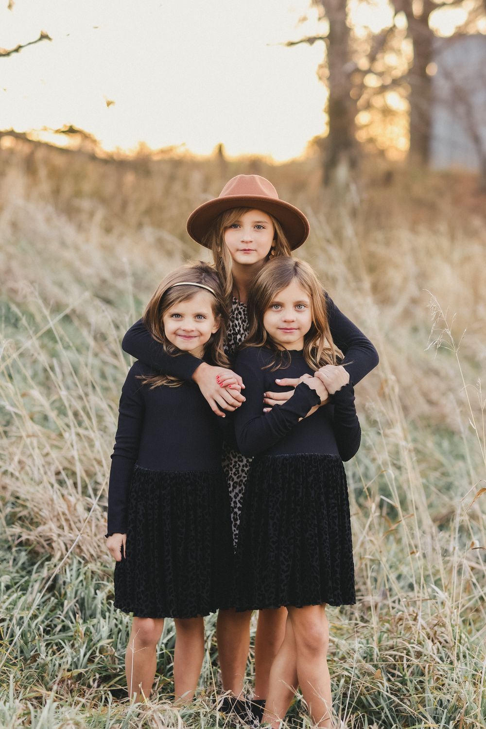 Three people in black dresses and brown hat pose in sunlit field.