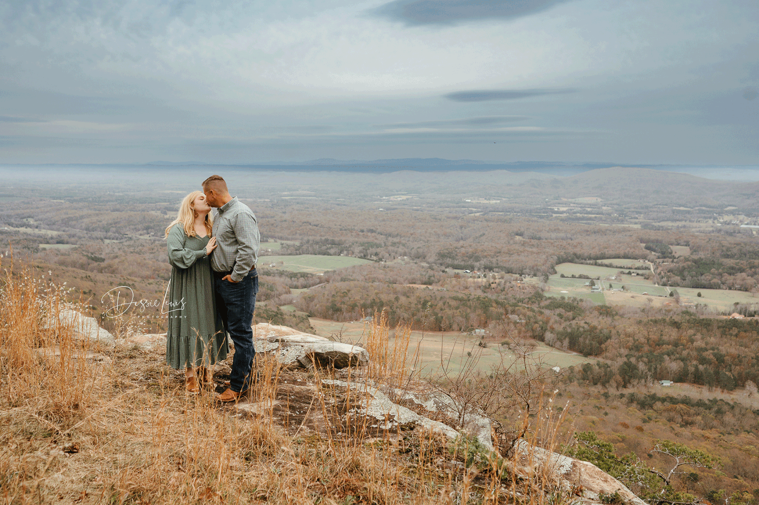 A couple shares an intimate moment overlooking a vast valley landscape on a cloudy day.