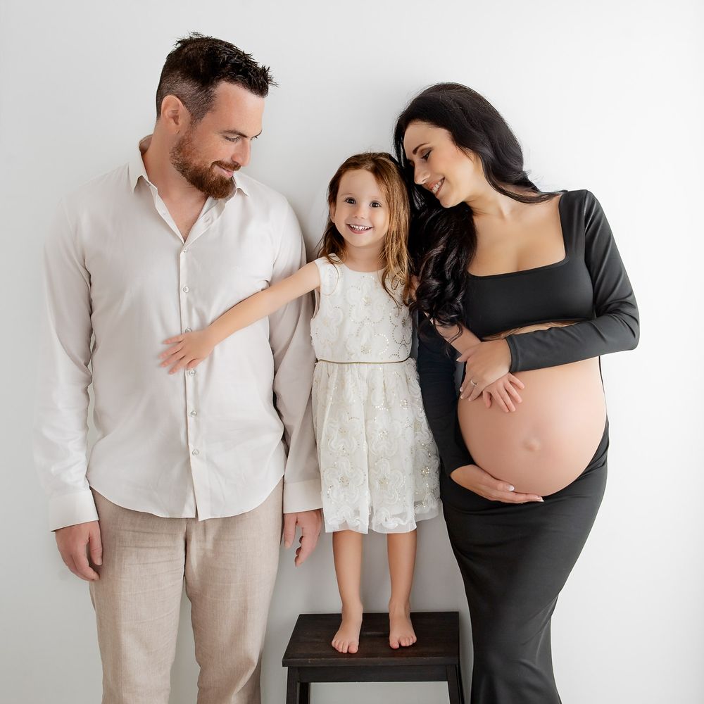 A family poses together against a white backdrop with a young girl standing on a stool and an expectant mother in black.