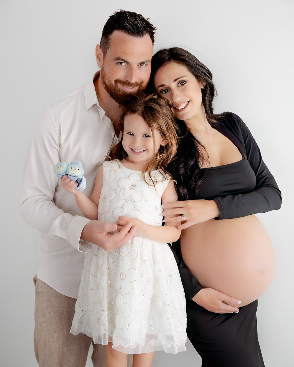 An intimate family portrait against a white background showing a young girl in white dress and expecting mother in black.