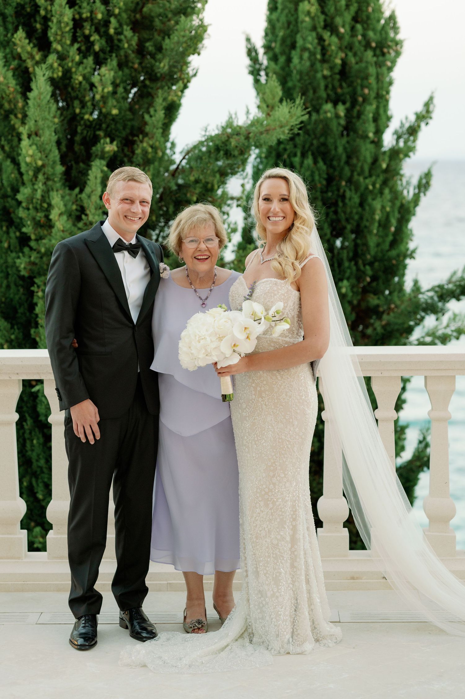 Elegant wedding portrait on a coastal terrace with towering cypress trees in the background.