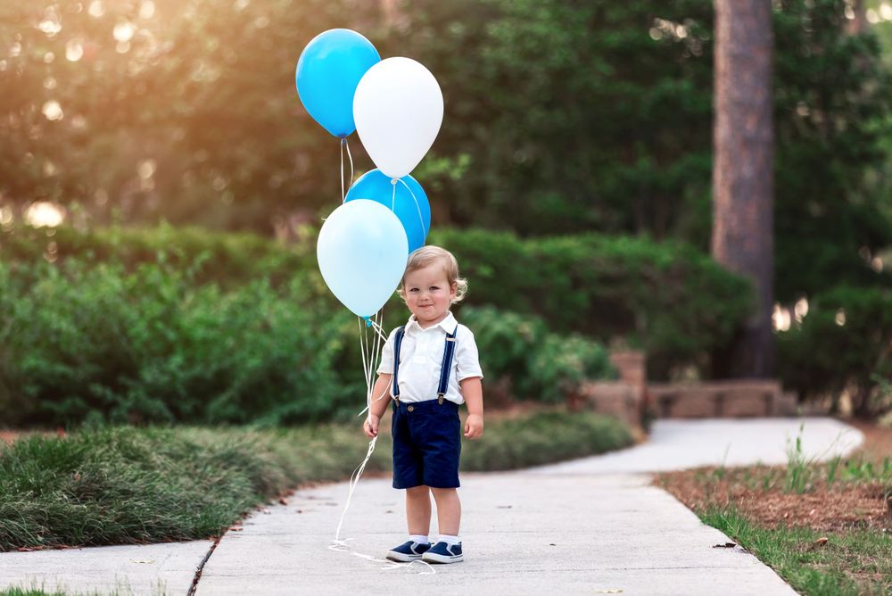 Blue and white balloons float against a golden sunset in a wooded park setting.