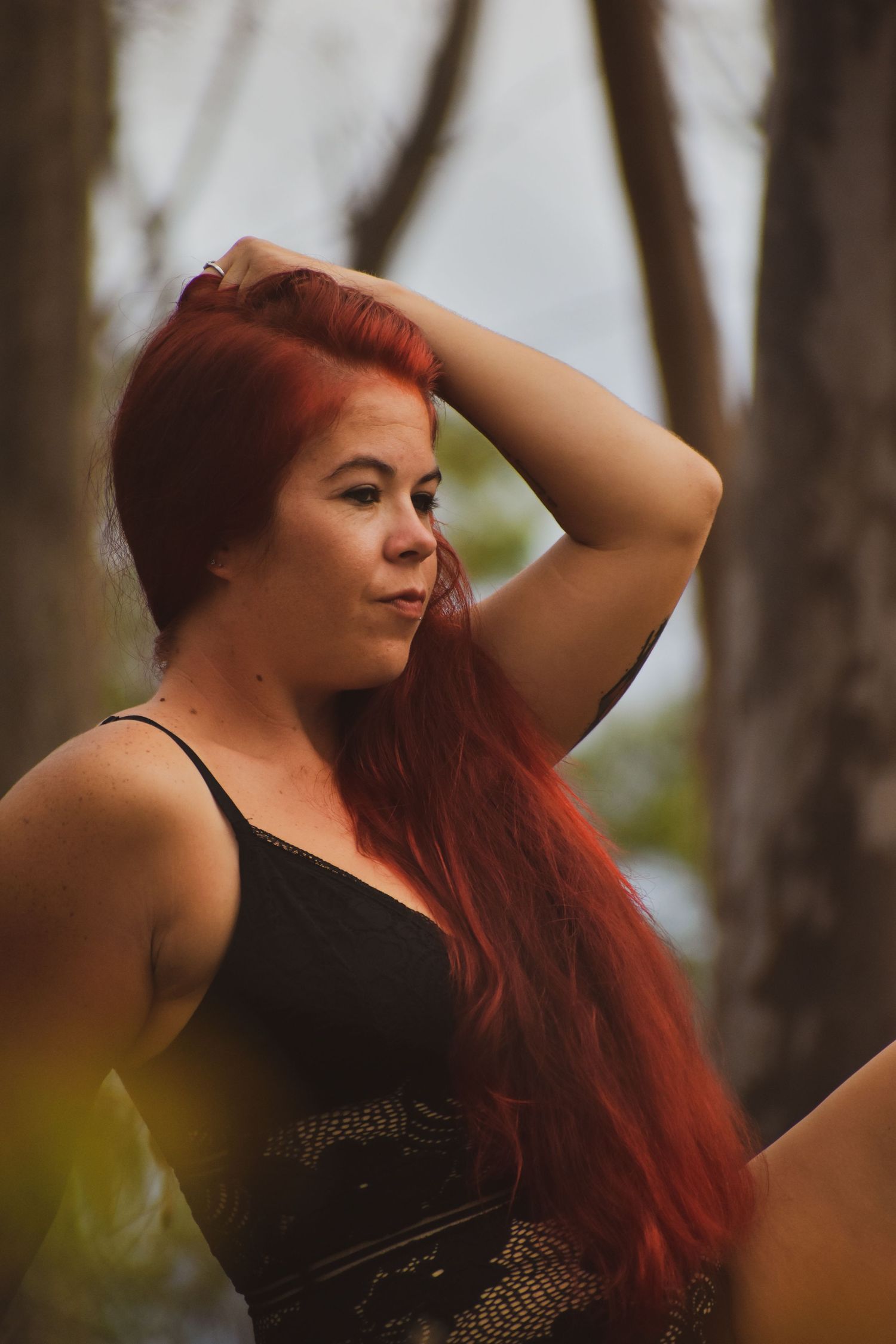 A woman with vibrant red hair poses outdoors in a black top against a natural forest backdrop.