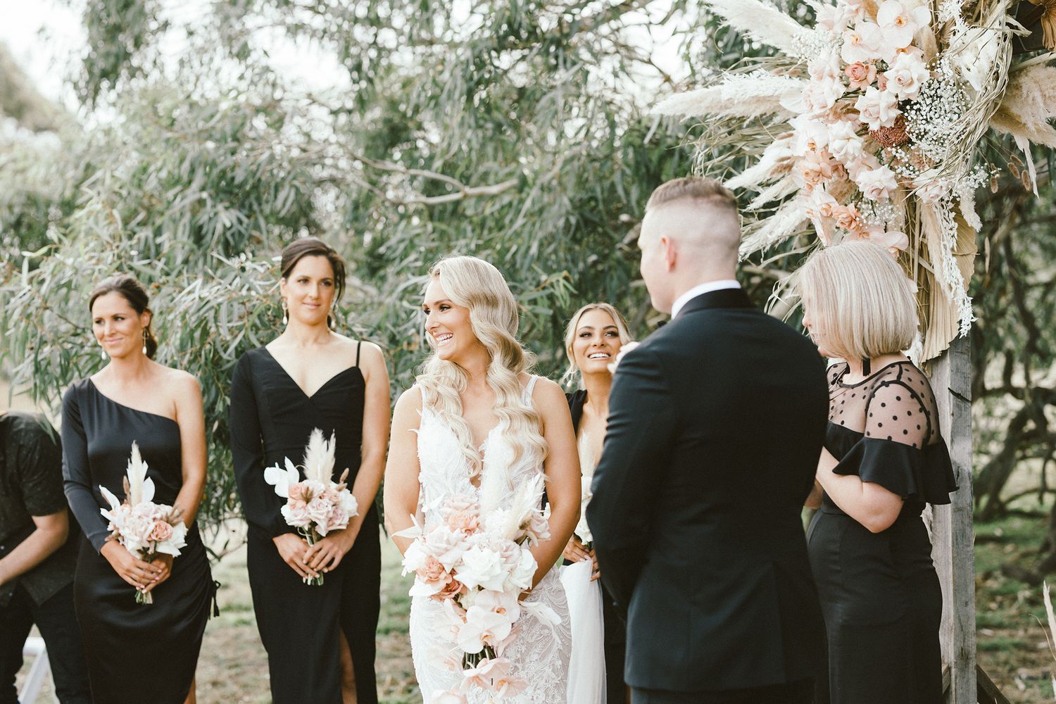 Wedding ceremony with bridesmaids in black dresses holding white floral bouquets.