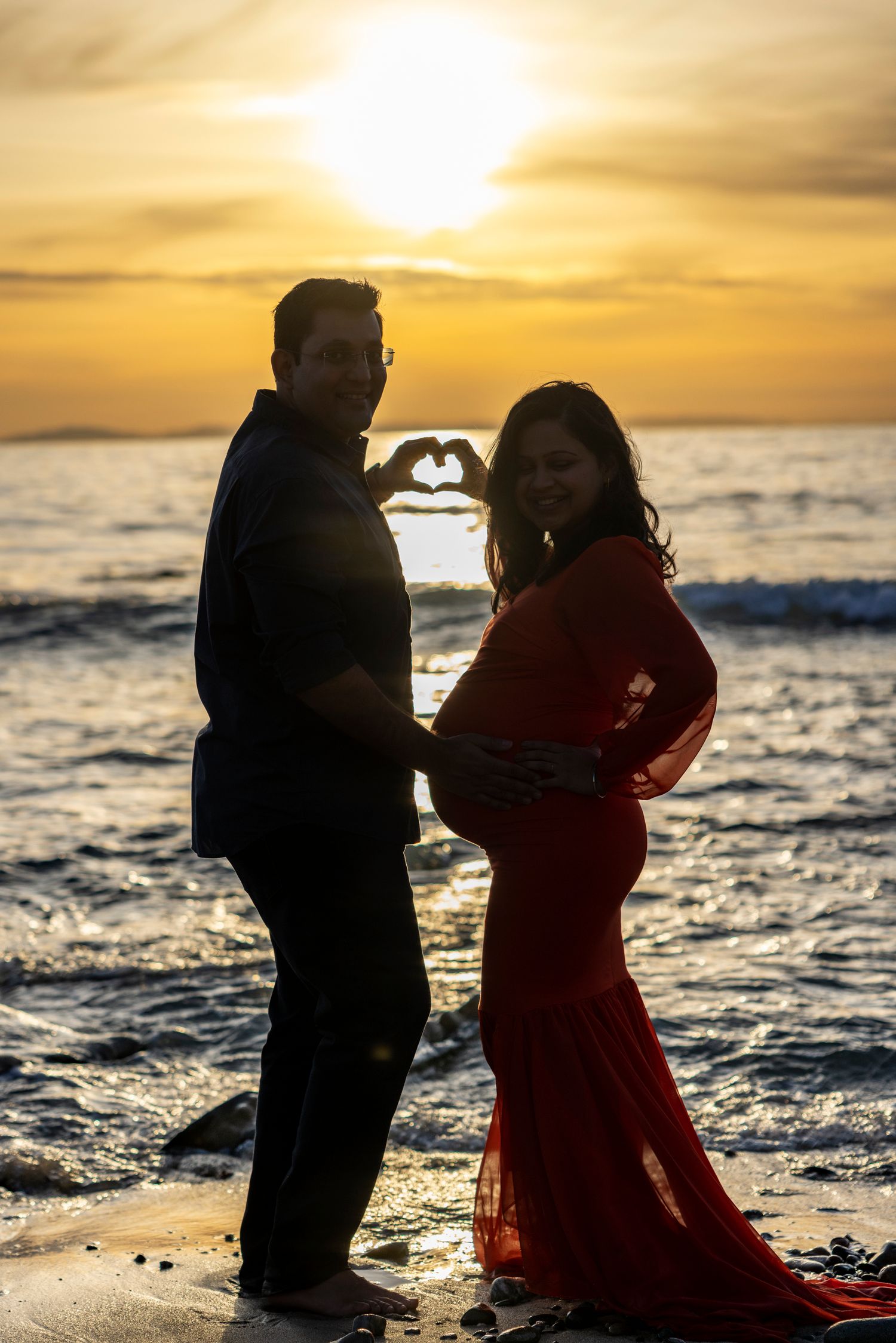 Beach maternity photoshoot with Couple creating a heart shape with hands at sunset beach, woman in red maternity gown.