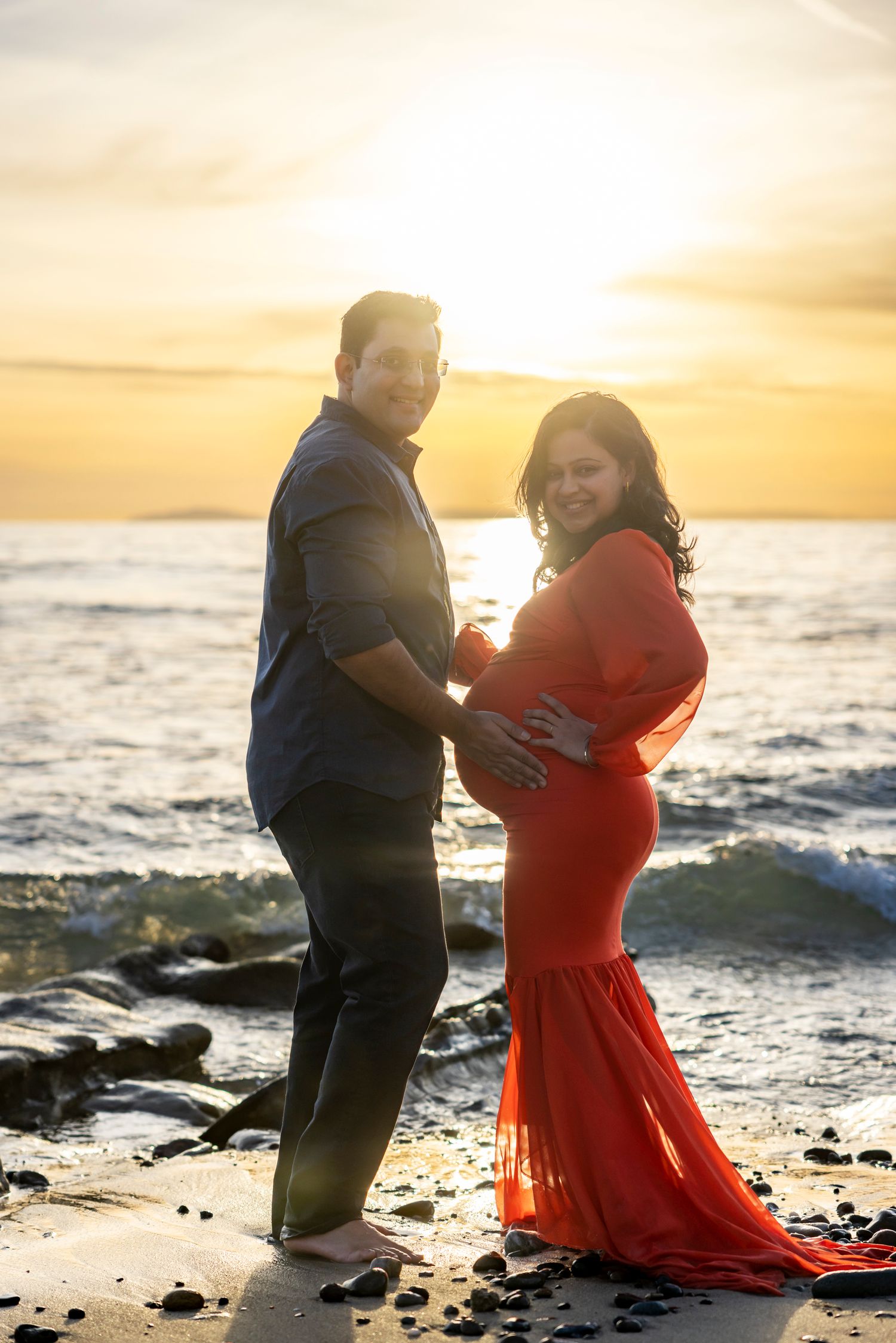 Beach maternity photoshoot with Couple posing romantically at sunset on the beach, woman in red maternity gown embracing her bump.