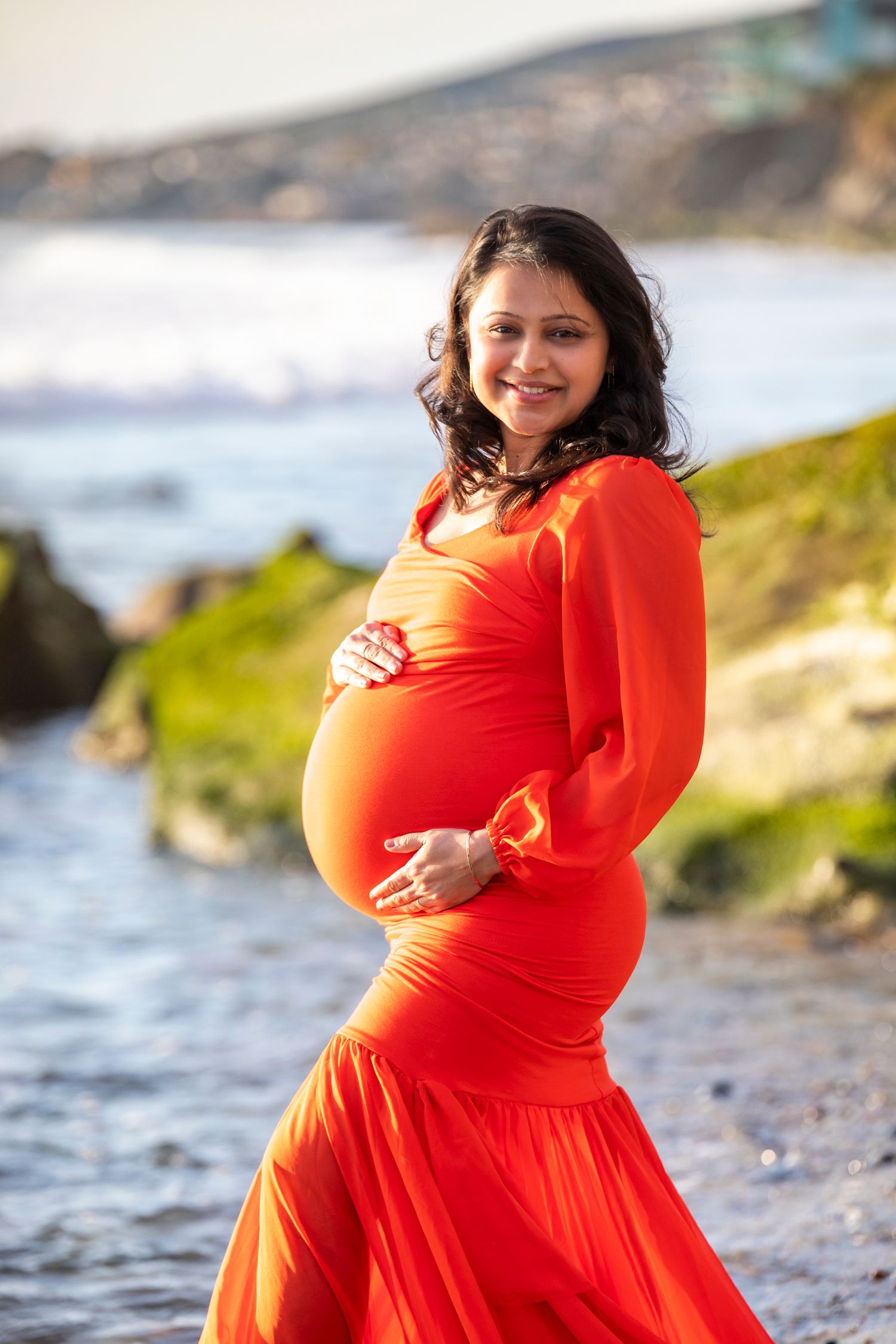 Beach maternity photoshoot with Smiling pregnant woman in flowing red dress holding her baby bump at a scenic ocean beach.