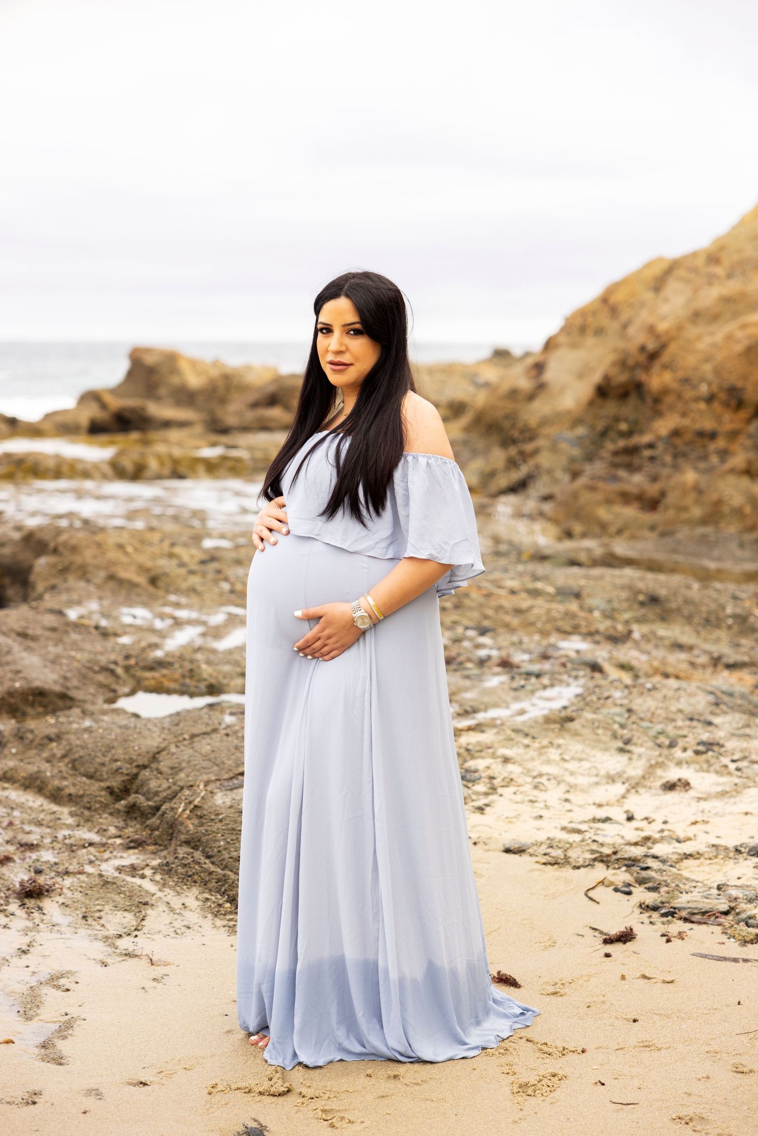 Beach maternity photoshoot with Pregnant woman in flowing light blue maternity gown posing on a rocky beach with ocean backdrop.