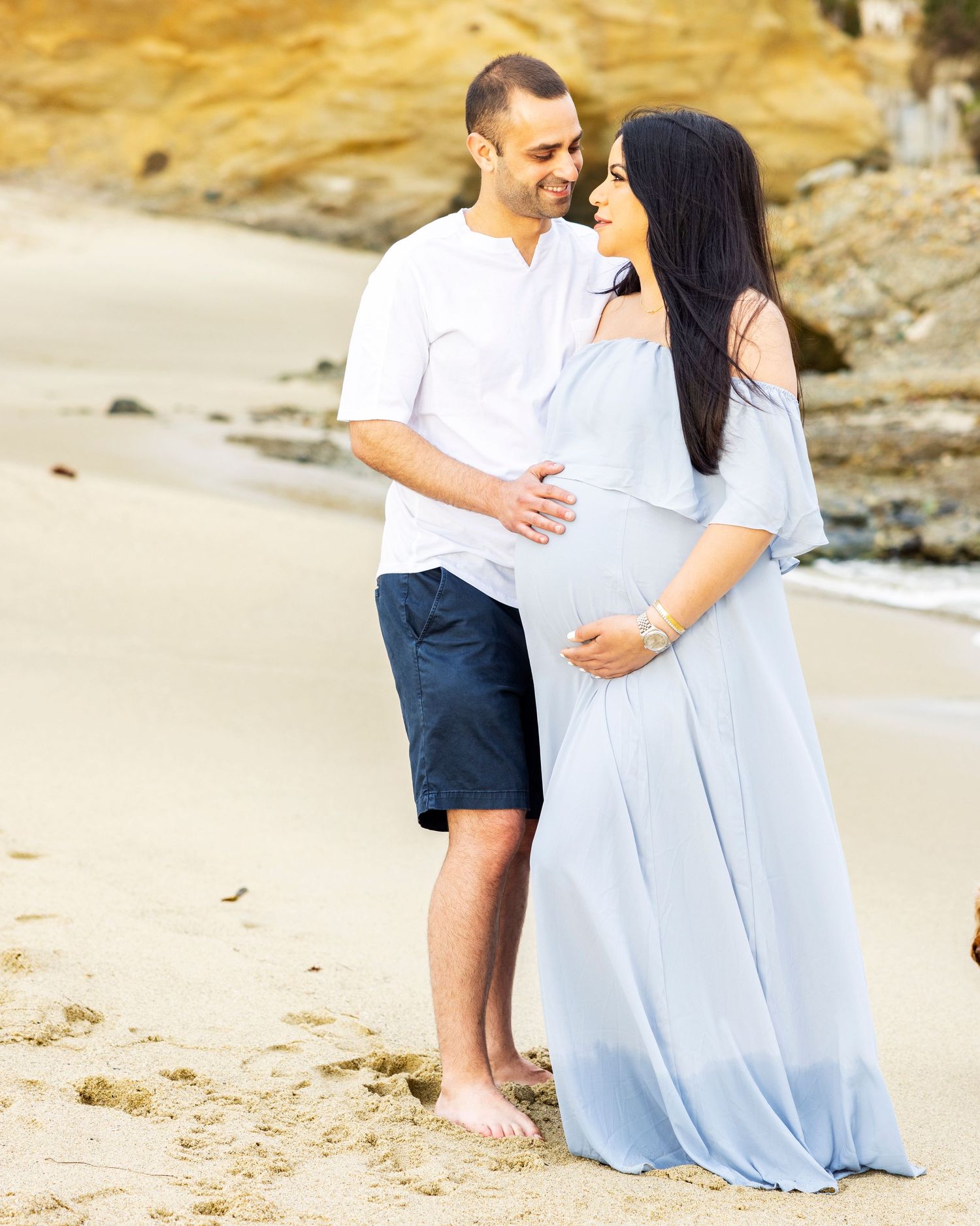 Beach maternity photoshoot with Couple sharing a tender moment on a sandy beach, woman wearing a light blue maternity gown.