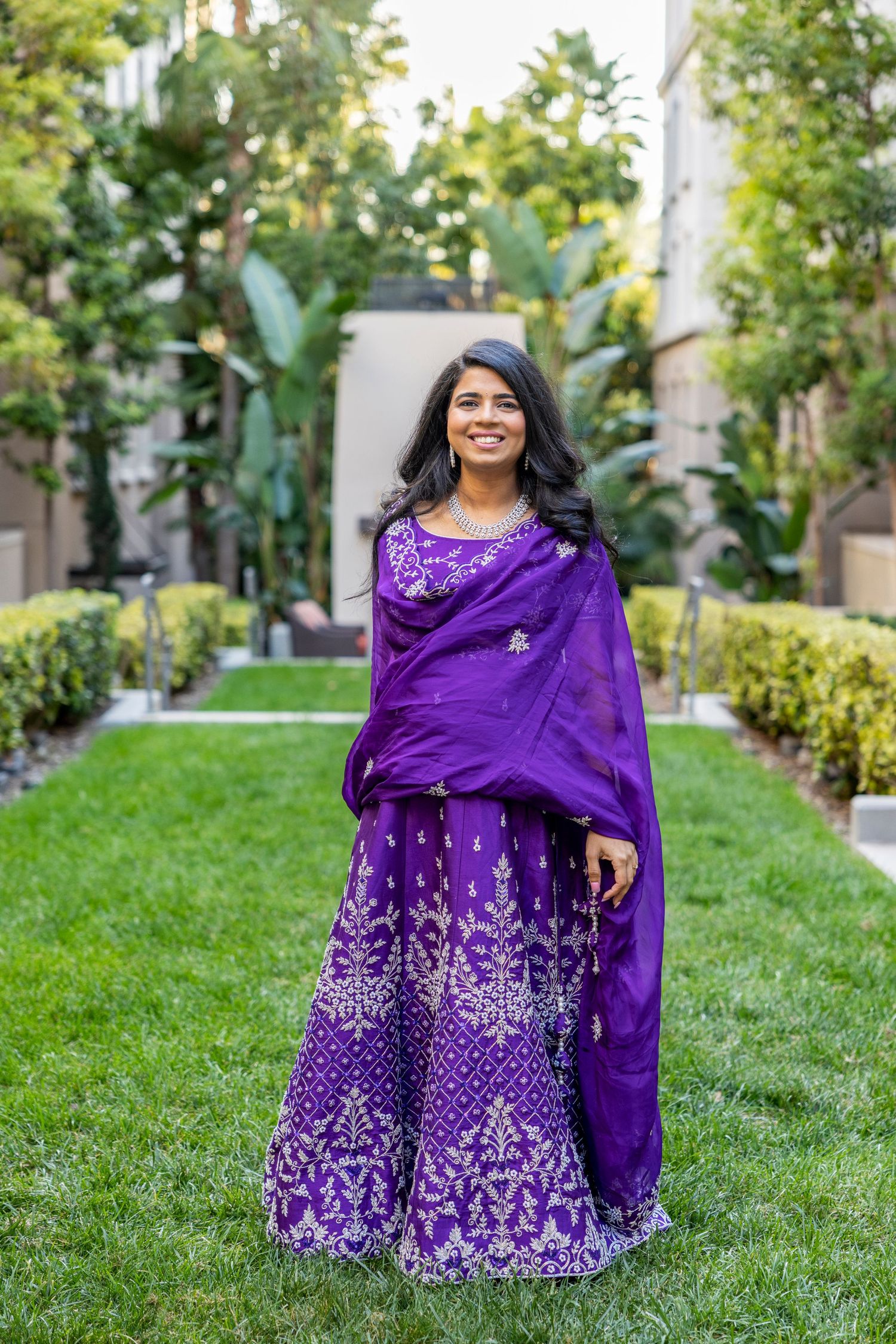 Smiling woman in ornate purple Indian lehenga standing on a lush green lawn outdoors.