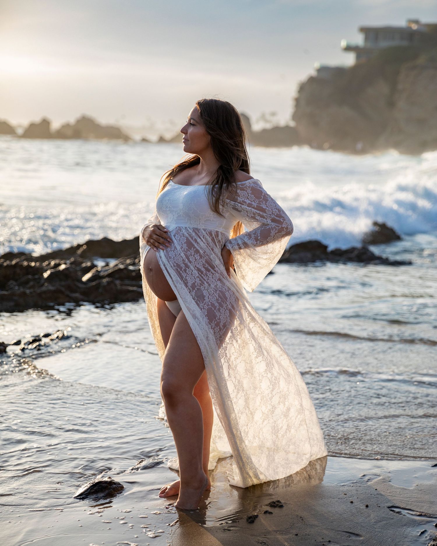 Beach maternity photoshoot with Pregnant woman in white lace maternity gown standing on beach at sunset with ocean waves.