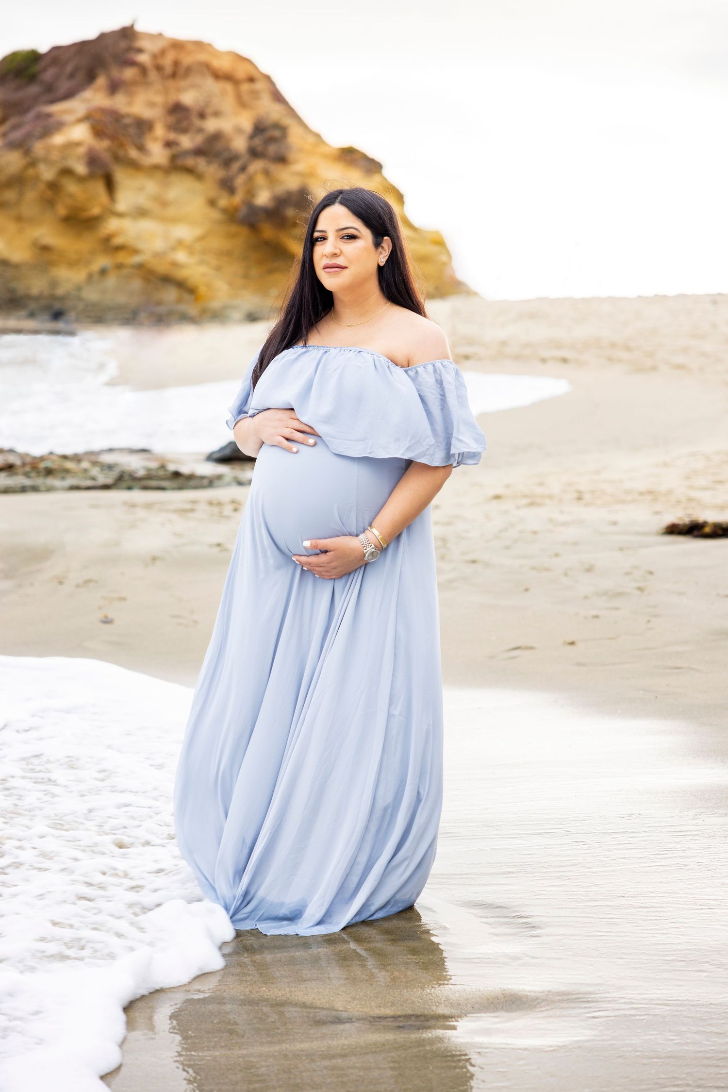 Beach maternity photoshoot with Pregnant woman in light blue off-shoulder maternity gown standing gracefully at the beach shoreline.