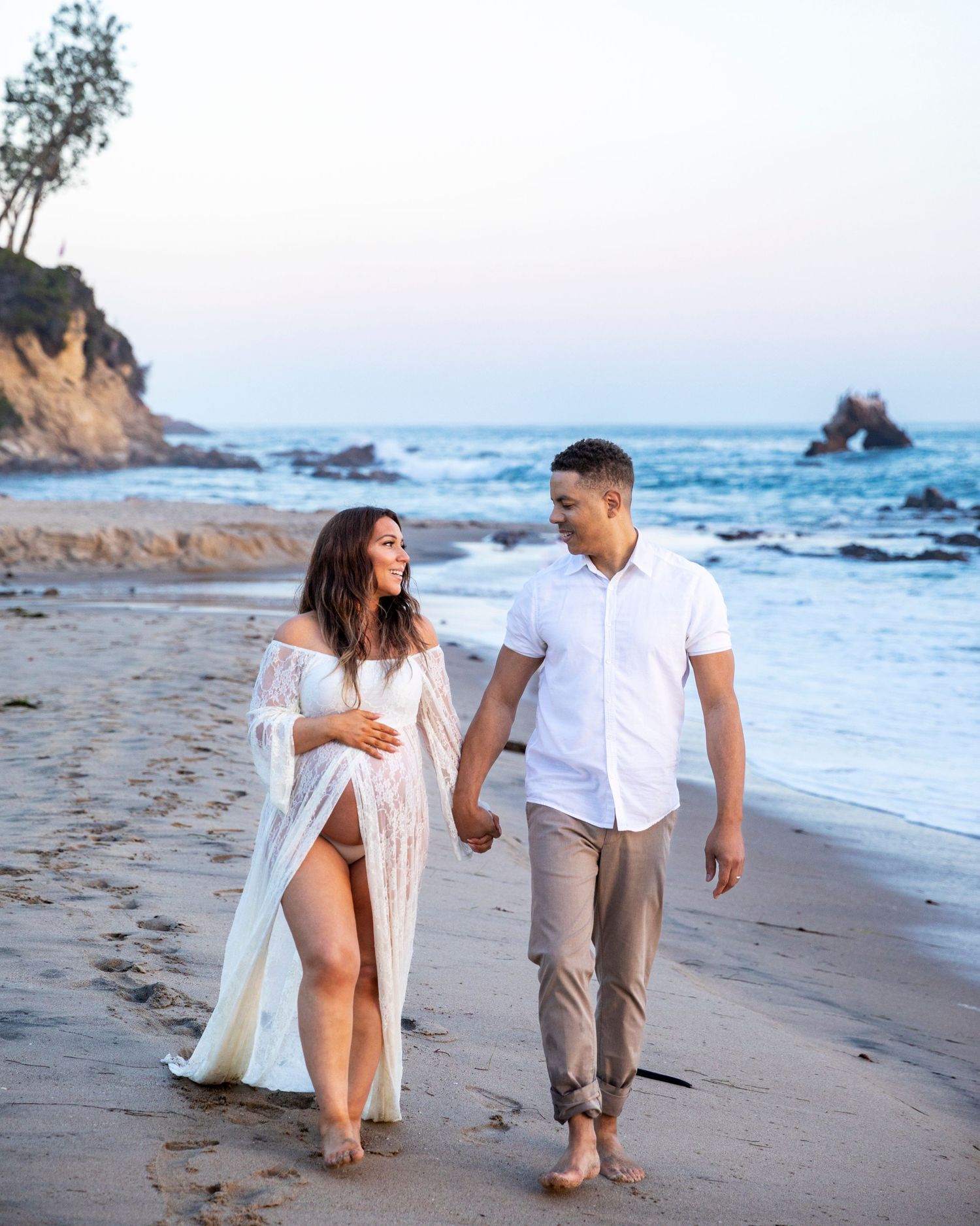 Beach maternity photoshoot with Expecting couple walking hand in hand on sandy beach at dusk in white outfits.