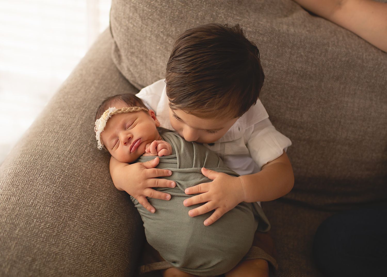 Toddler lovingly embraces newborn sibling while resting together on gray couch in warm lighting.