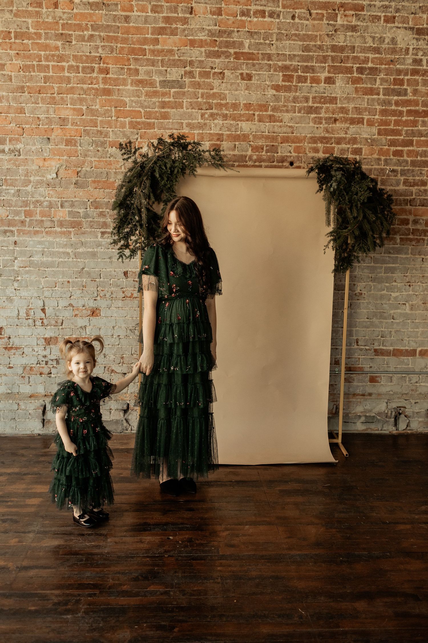 Mother and daughter wearing matching green dresses pose against exposed brick wall with floral arch.
