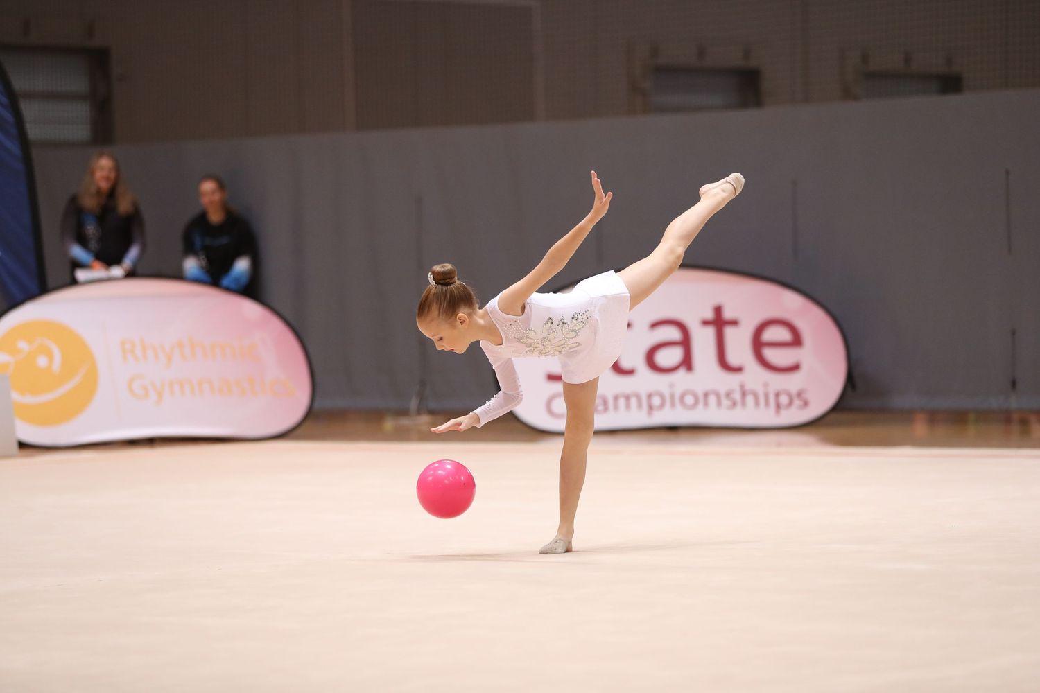 Rhythmic gymnast in white leotard performs graceful move with red ball during State Championships.