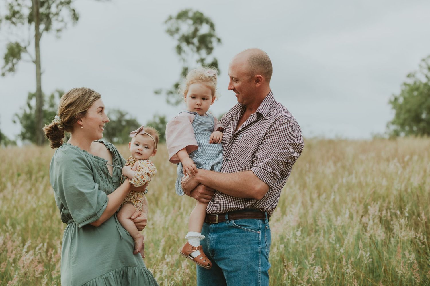 Smiling parents hold two young daughters in tall grass field, sharing a joyful family moment outdoors.