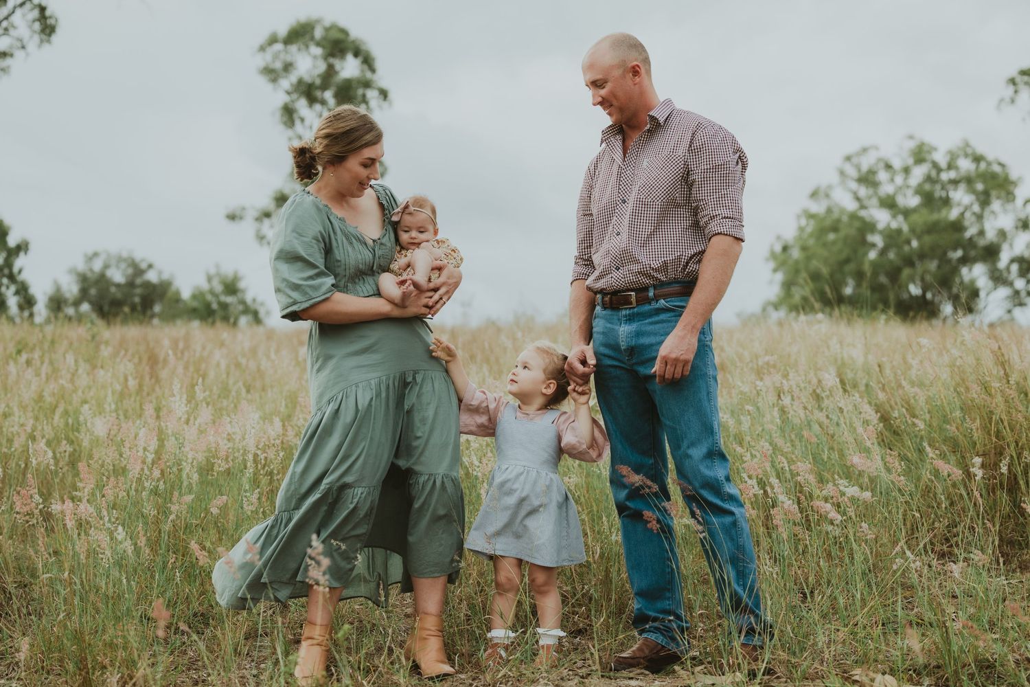 Family of four stands together in tall golden grass field, parents holding children on a cloudy day.