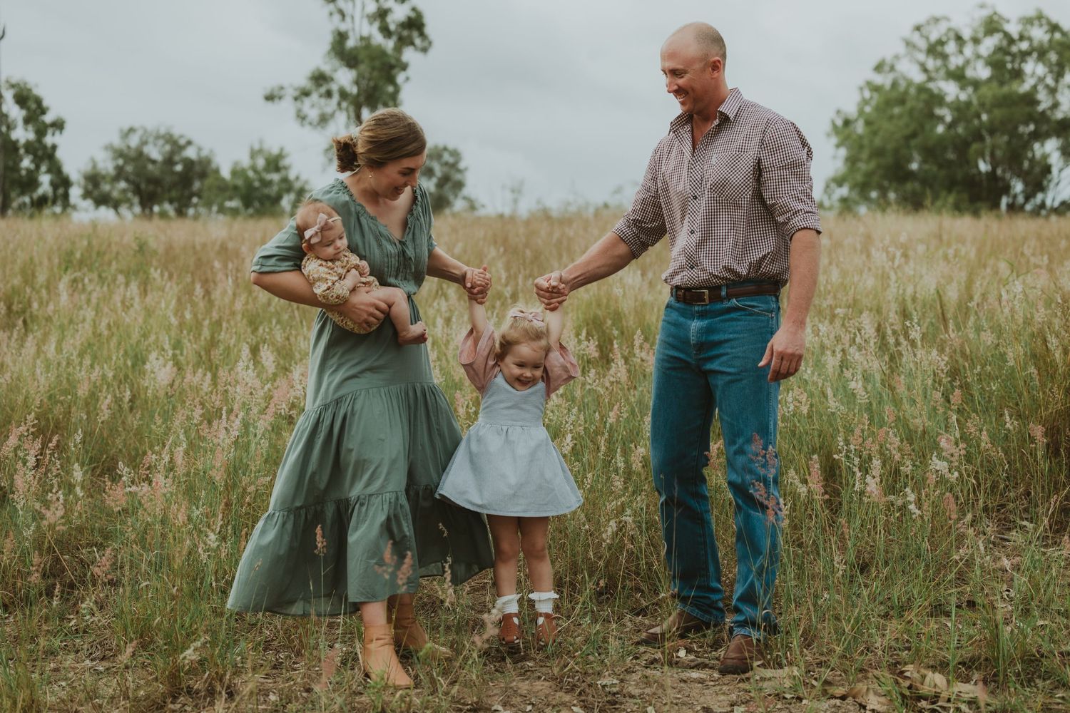 Parents walk with toddler daughter through tall grass field while mother carries newborn baby outdoors.