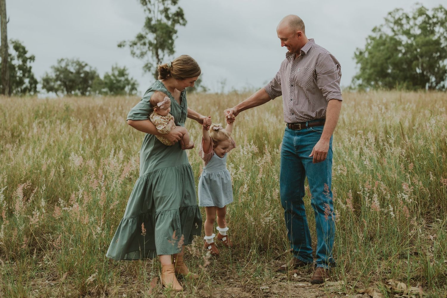 Parents guide toddler through golden grass field as mother carries infant during outdoor family photo session.