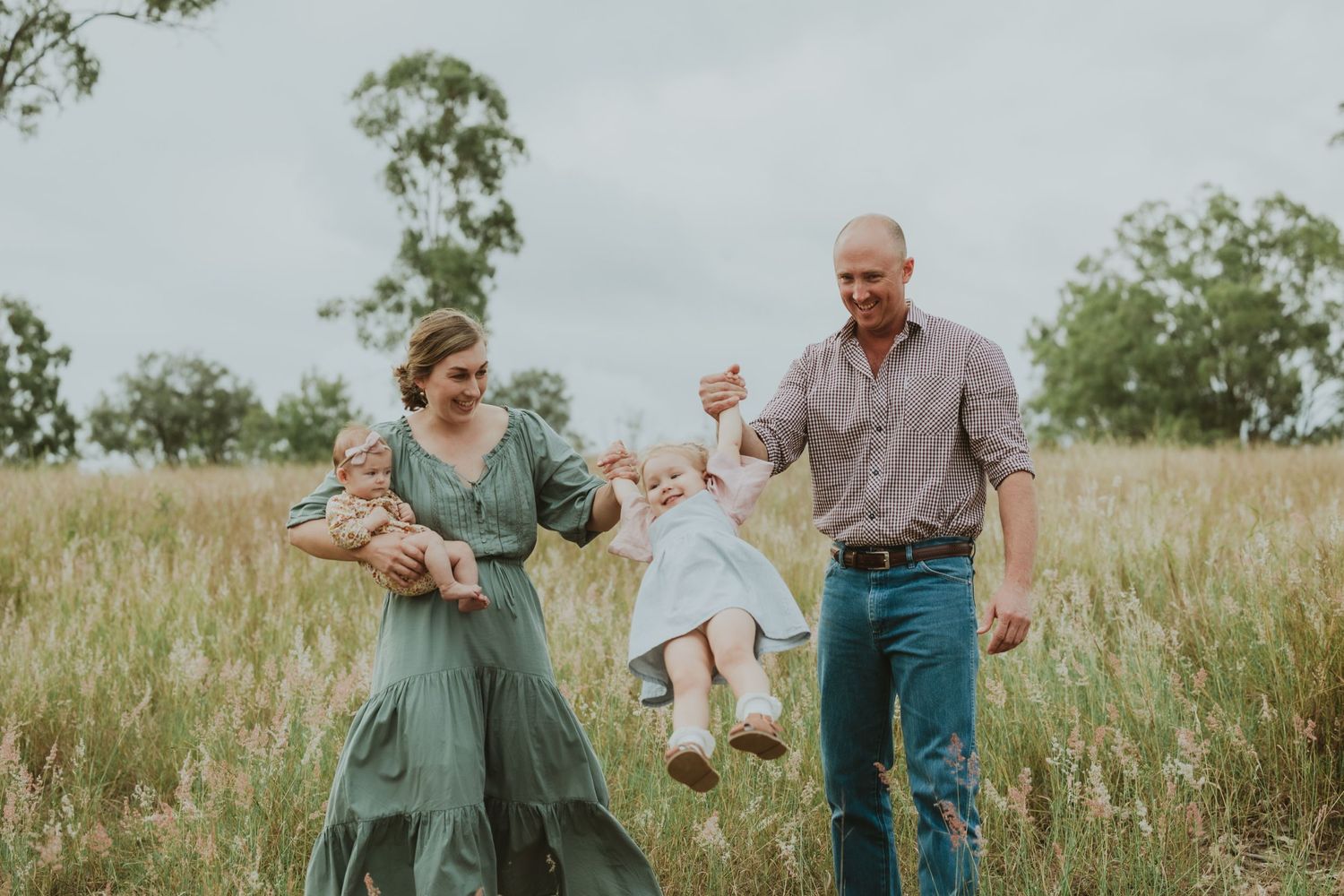 Joyful parents swing laughing toddler between them in golden grass field while mother holds newborn baby.