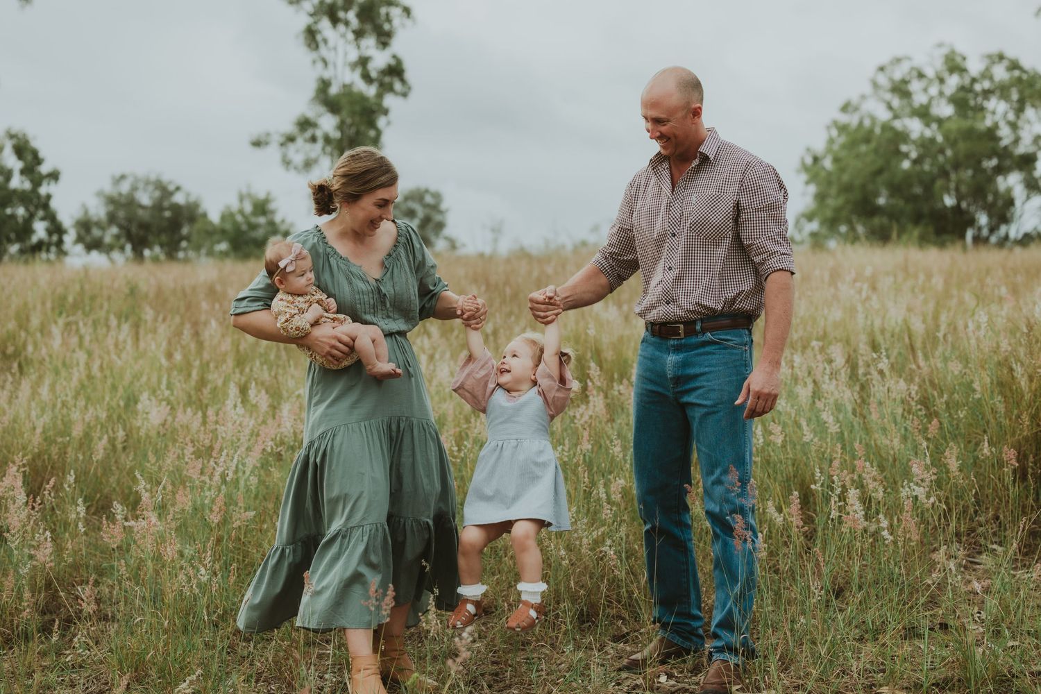 Parents hold hands with toddler daughter walking through tall golden grass field with newborn baby.