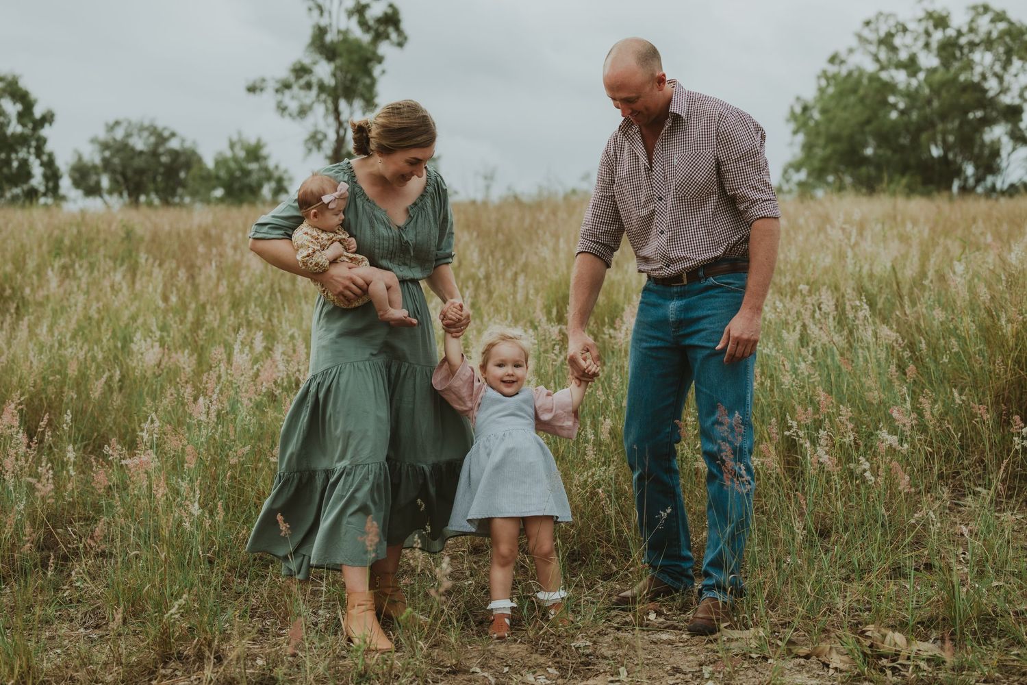 Parents walk hand-in-hand with toddler daughter through wildflower field while mother cradles newborn baby.