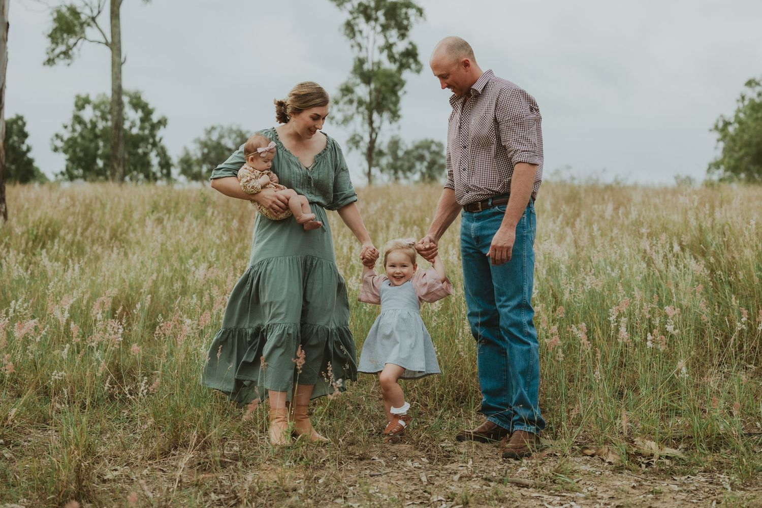 Family of four walking through golden grass field, parents holding toddler's hands while mom carries newborn baby.