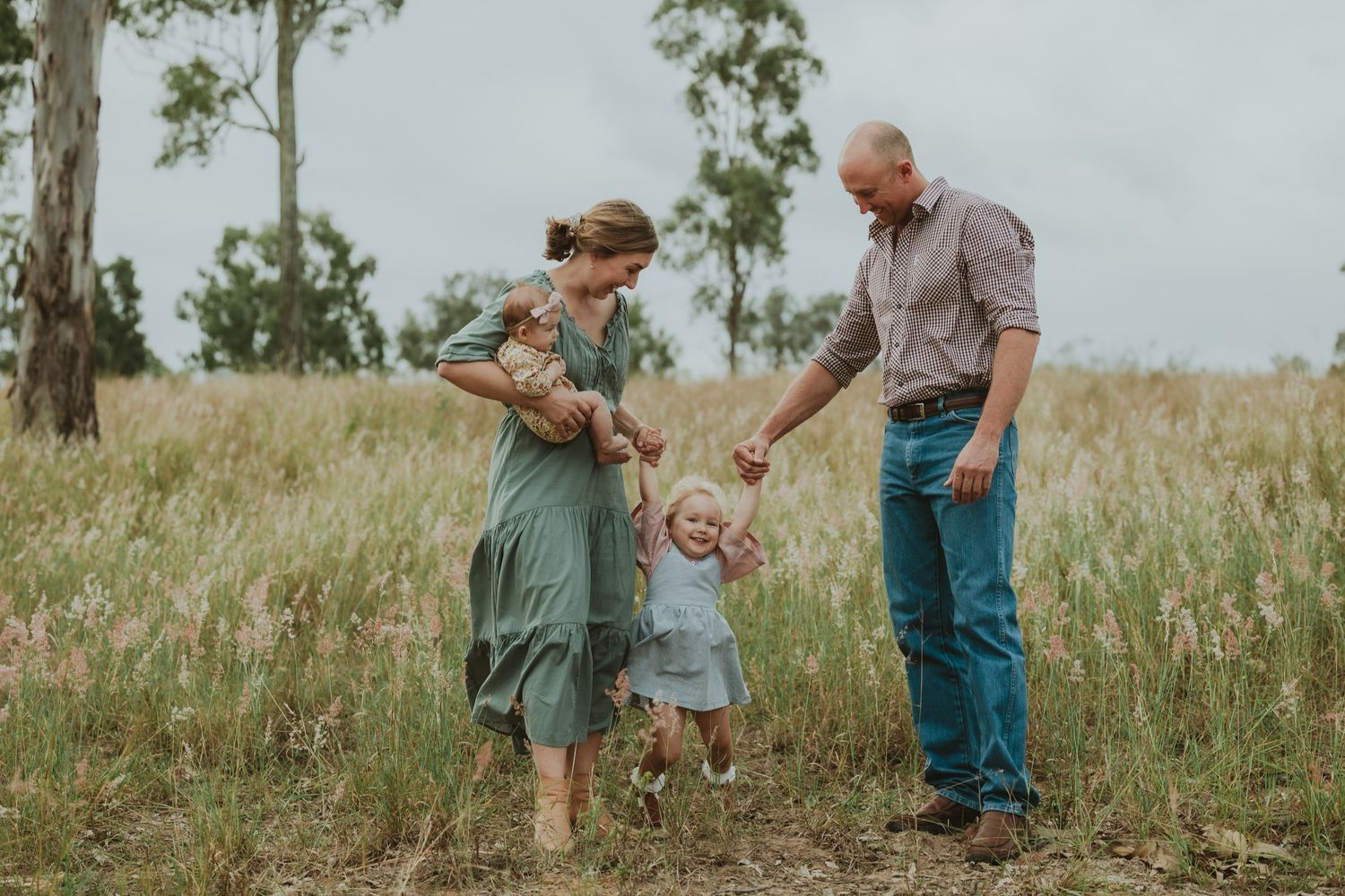 Happy family with young child playing in tall grass meadow, parents swinging toddler between them outdoors.
